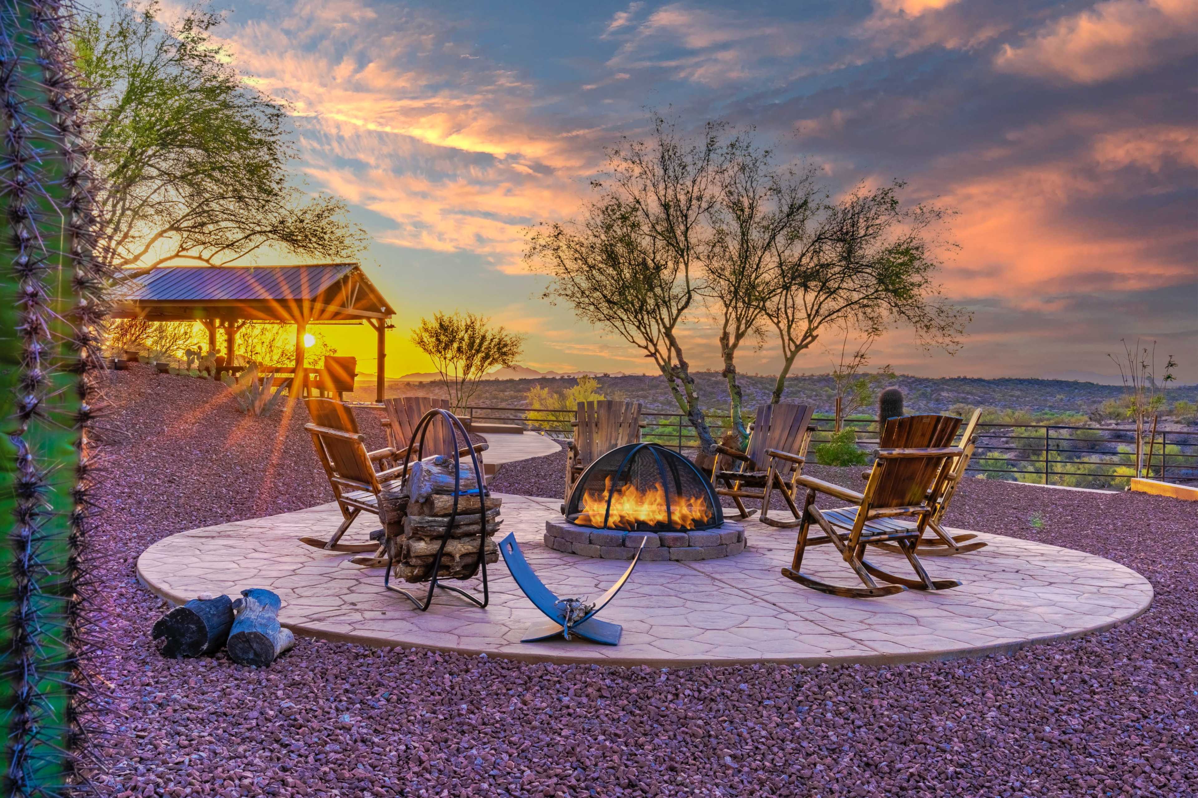 A circular fire pit surrounded by wooden chairs is set against a colorful sunset over a desert landscape.