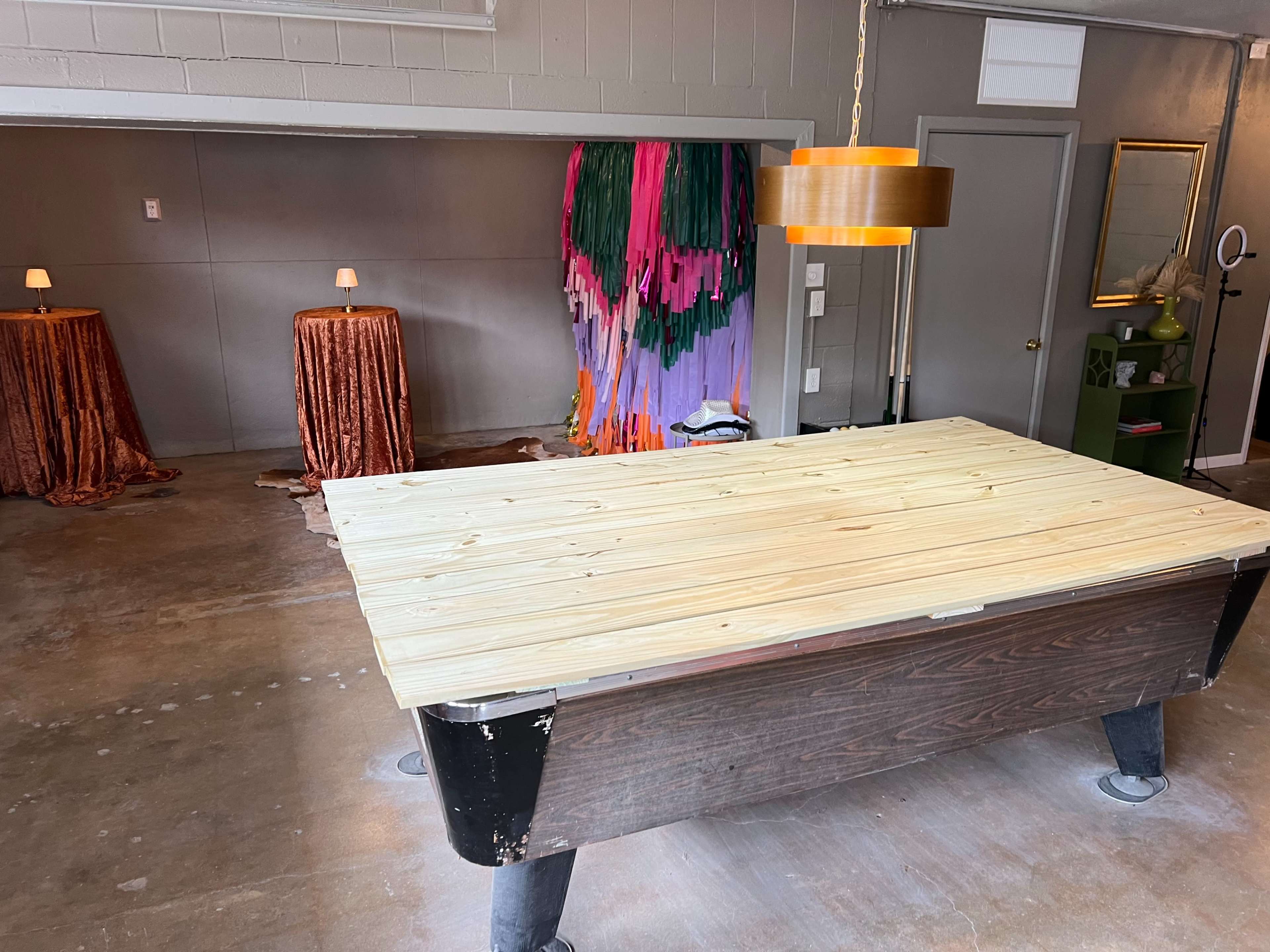The image shows a spacious room with a wooden pool table covered by a wooden plank, surrounded by two decorative tables with lamps and a colorful fringed backdrop.