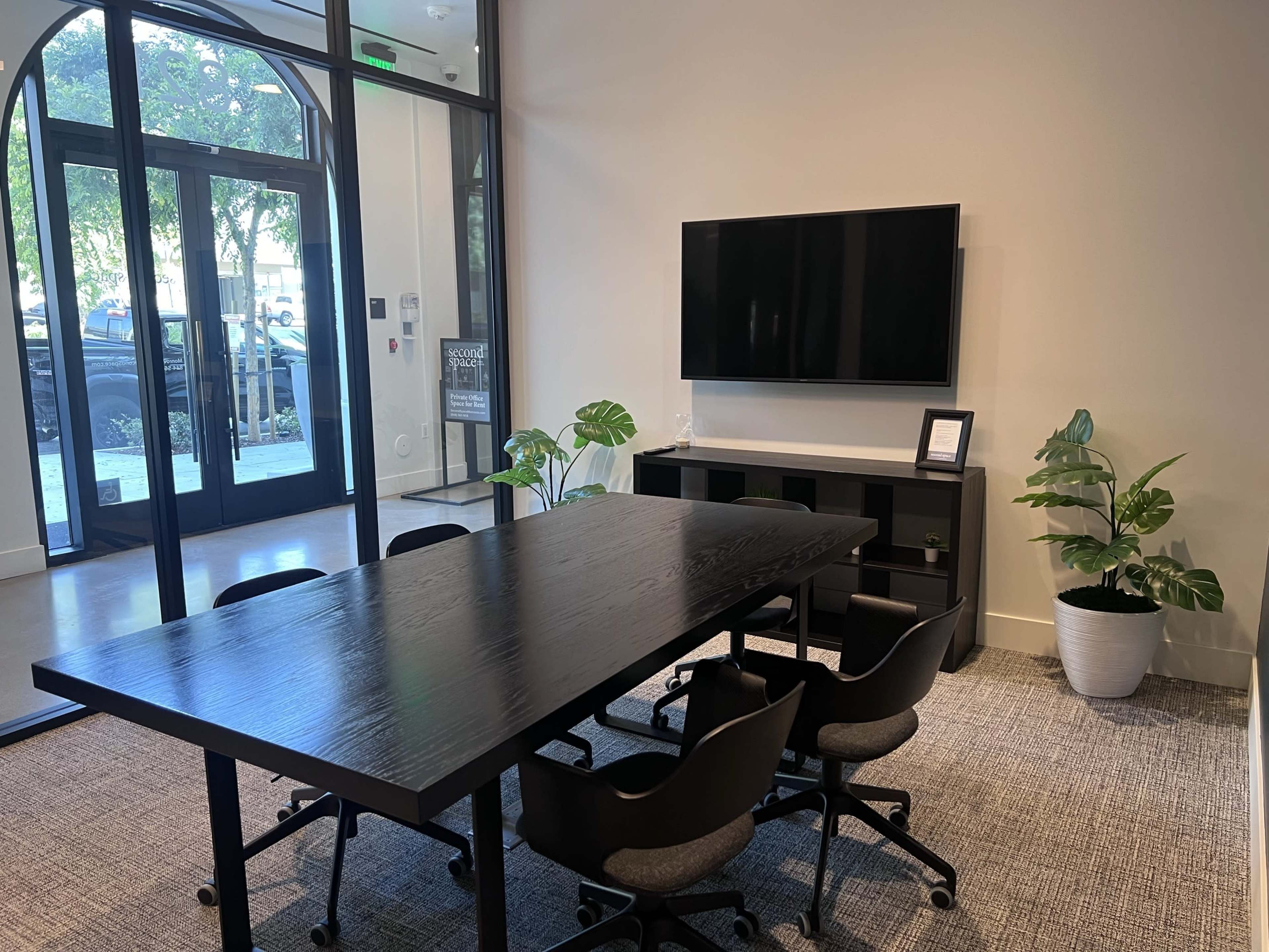 The image shows a modern conference room with a large black table, six black chairs, a wall-mounted television, and two potted plants.