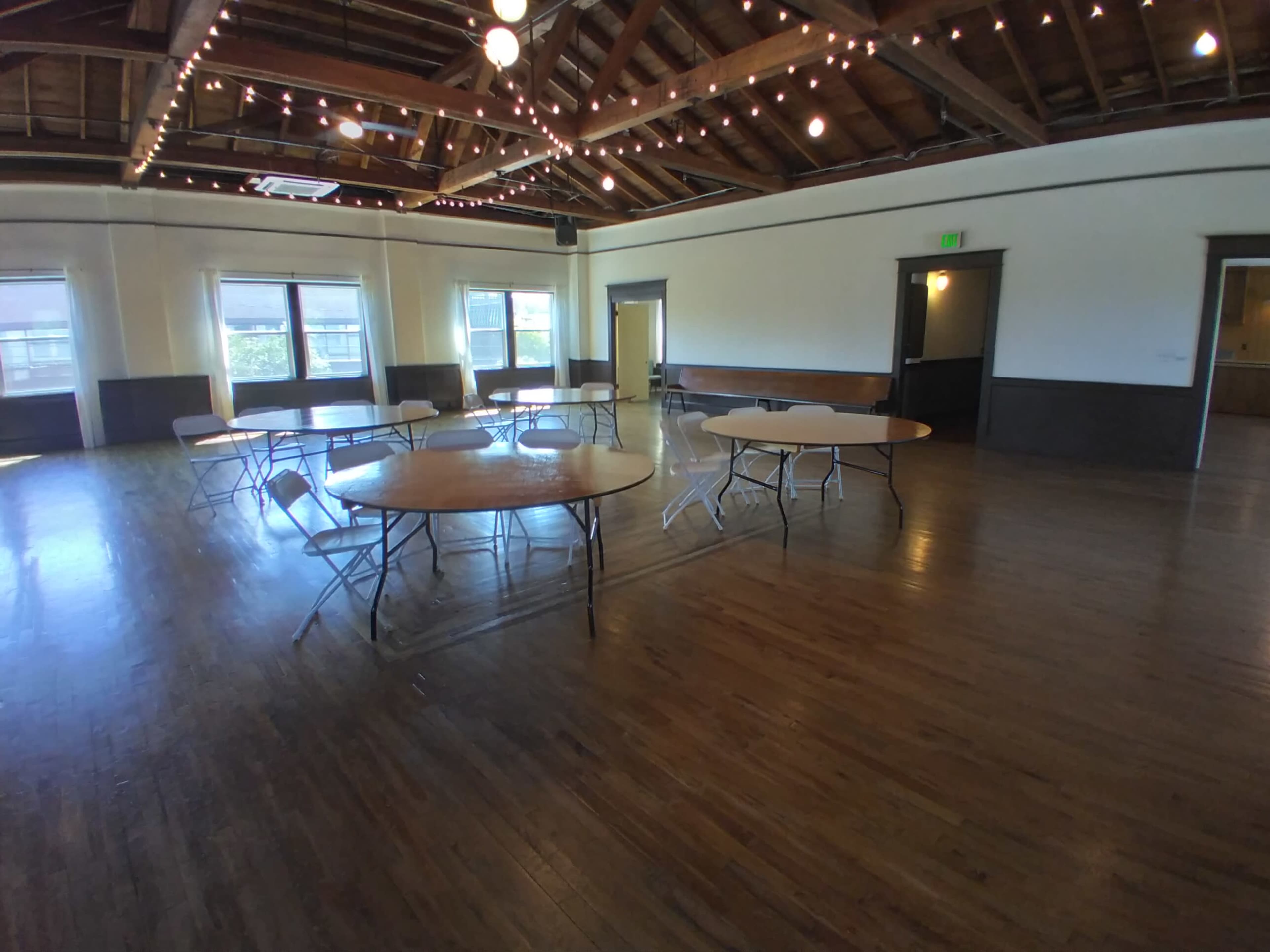 A spacious room features several round tables surrounded by folding chairs, with wooden flooring and exposed beams illuminated by string lights.