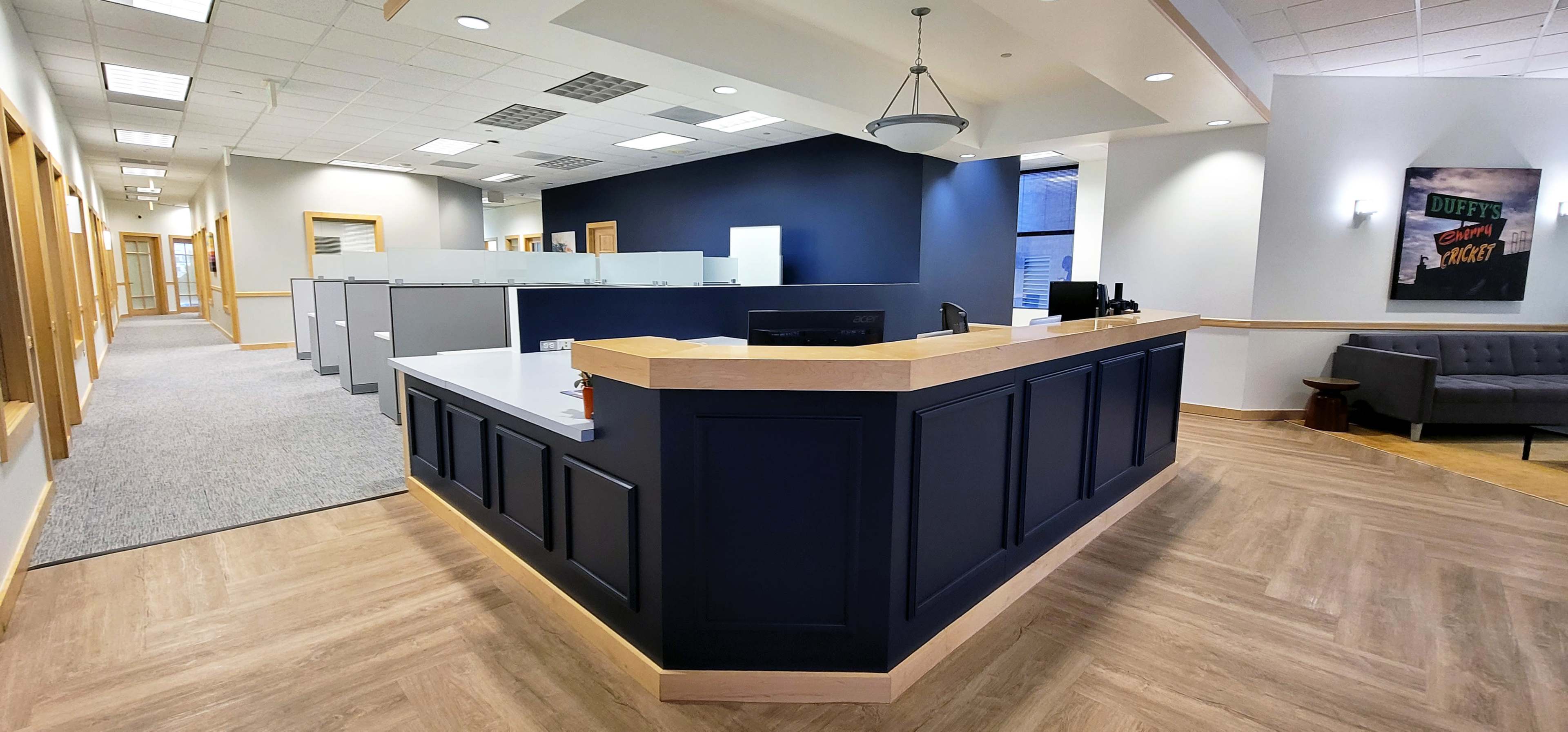 A modern office reception area featuring a wooden reception desk with gray cubicles in the background and a seating area on the side.