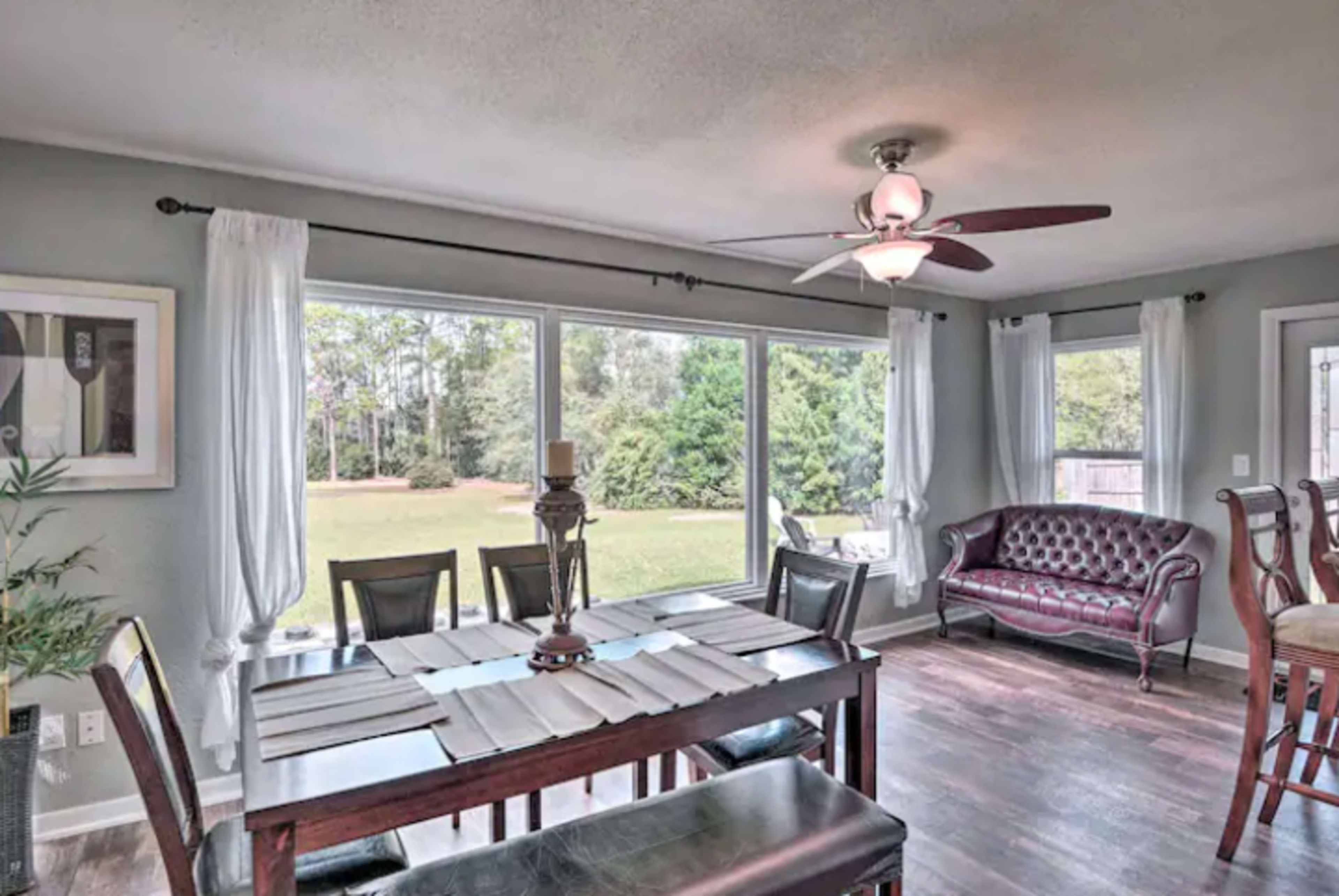 A dining area with a wooden table, eight chairs, and large windows overlooking a green outdoor space.