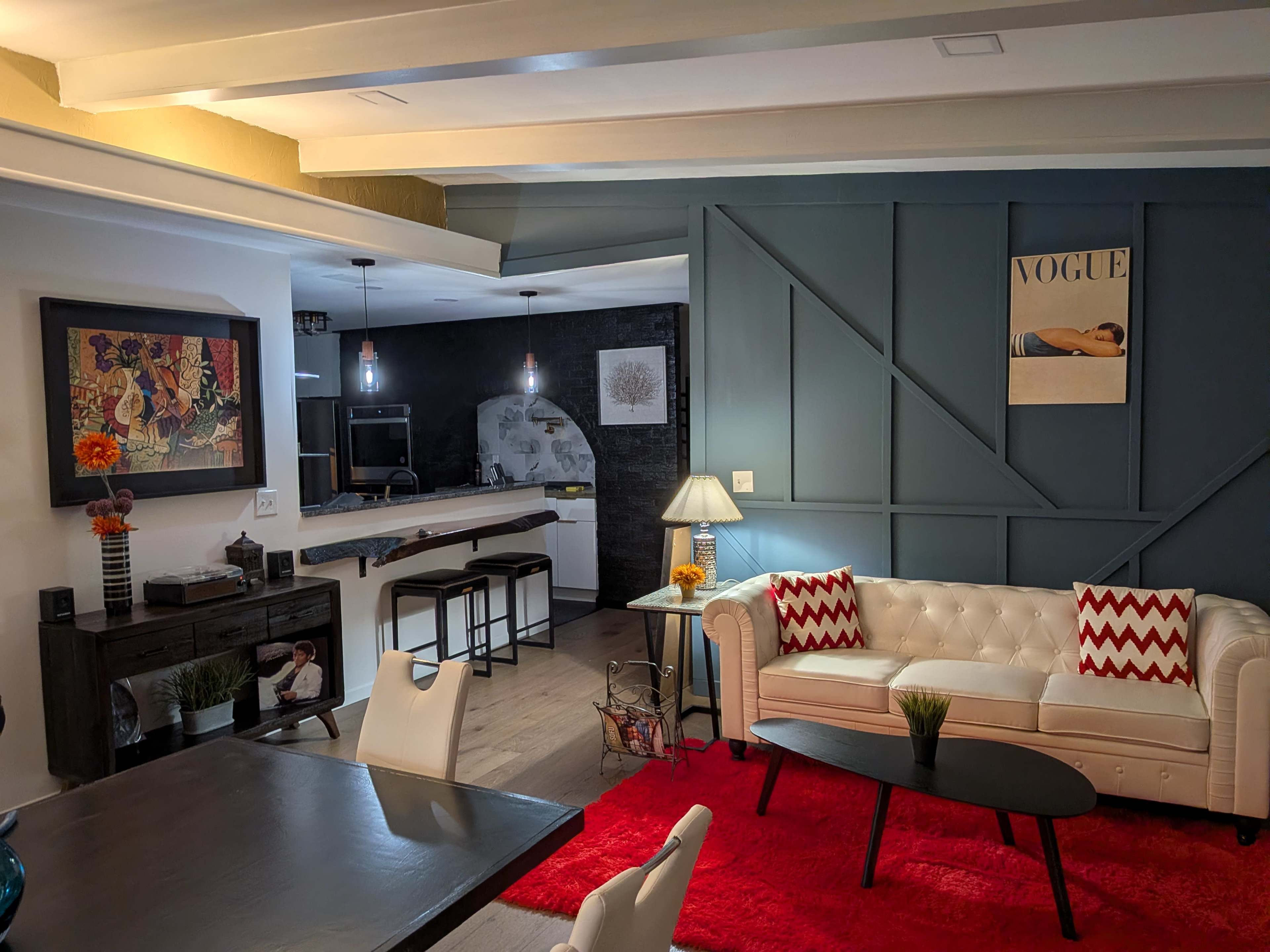 The image depicts a modern living room featuring a white leather sofa, a red rug, and a dining area with a table, adjacent to a kitchen with dark cabinetry and a Vogue magazine poster on the wall.