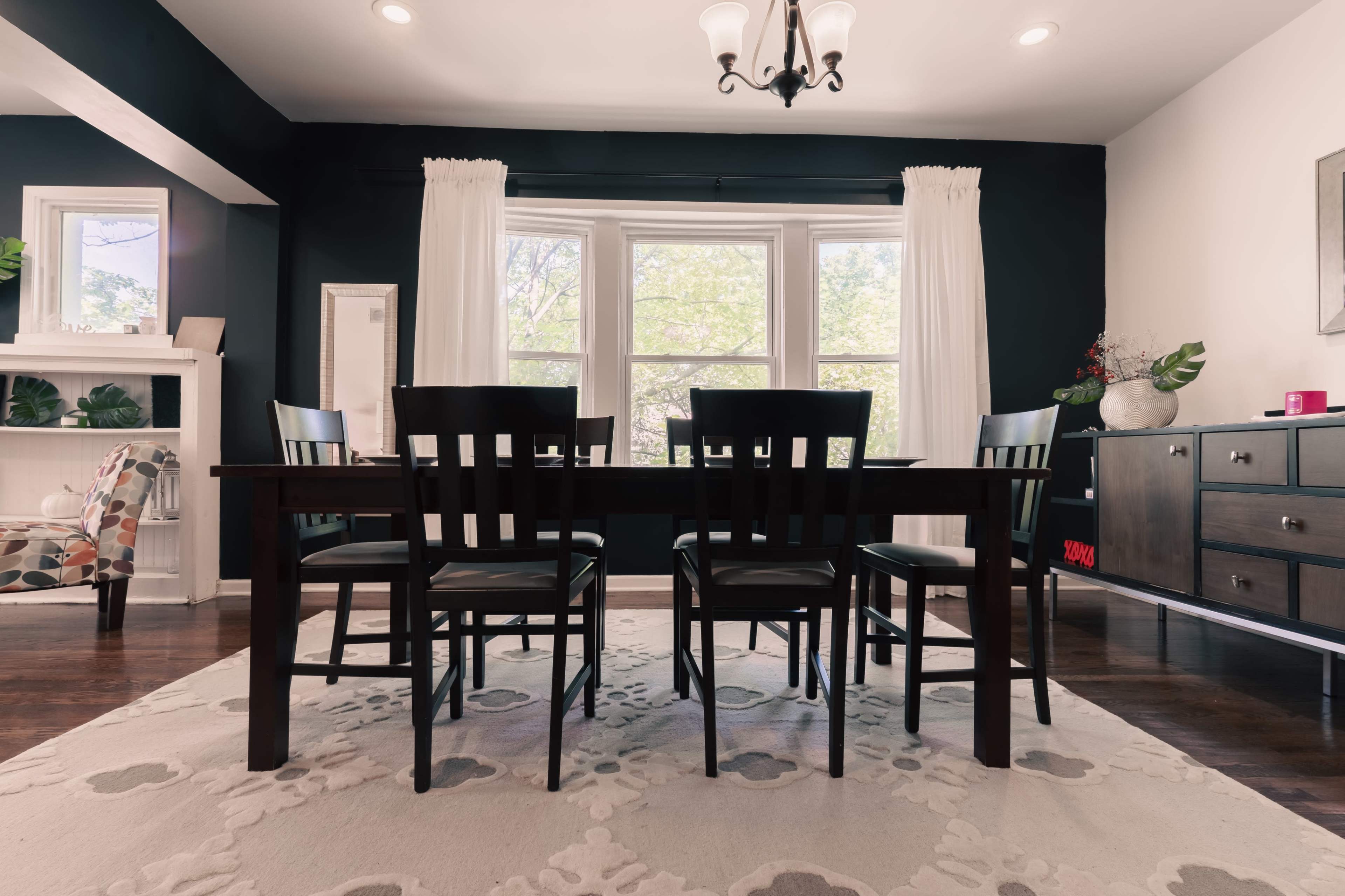 A dining room with a dark accent wall, a wooden table surrounded by four black chairs, and large windows letting in natural light.