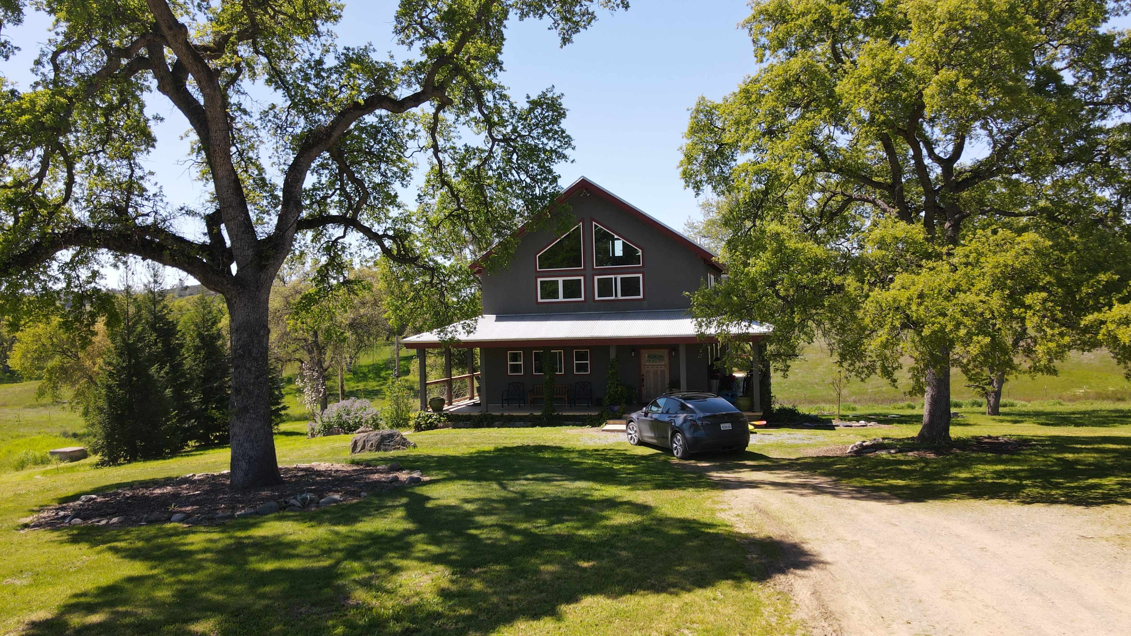 A two-story gray house with large windows and a porch is situated on a grassy landscape with trees and a car parked in front.