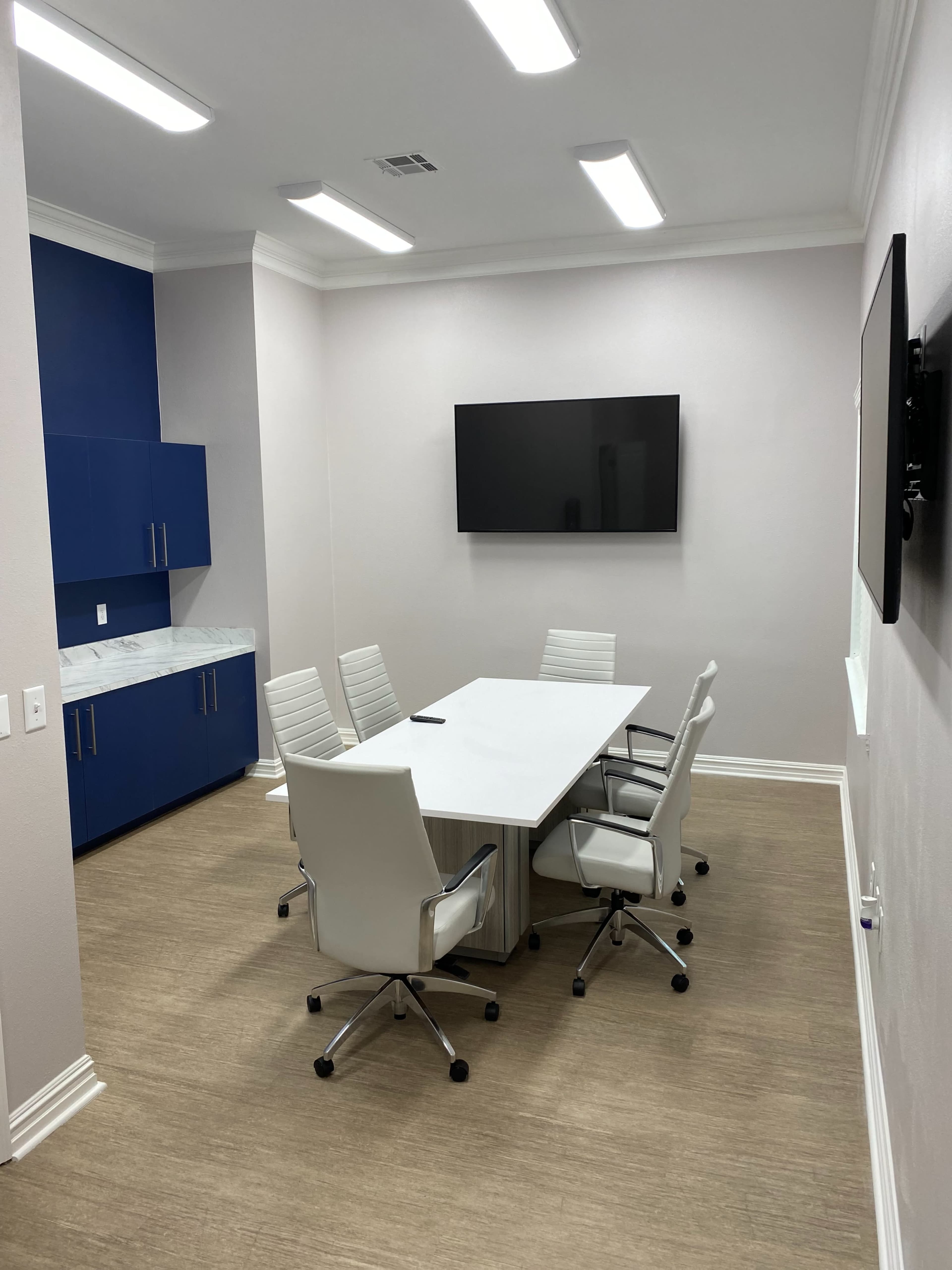 A small conference room with a rectangular table surrounded by six white chairs, blue cabinetry, and two wall-mounted TVs.