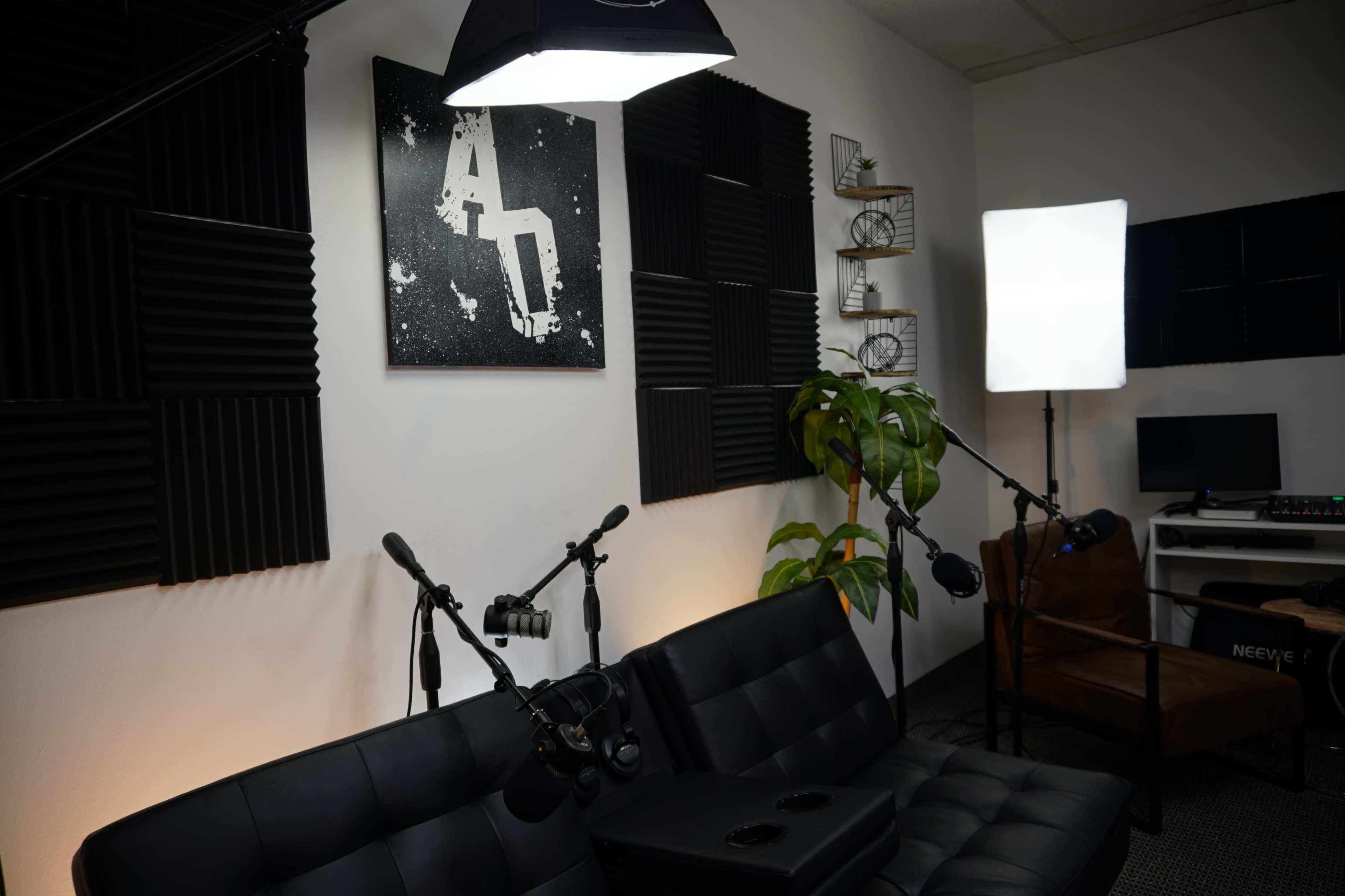 The image shows a room set up for recording or streaming, featuring acoustic foam panels on the walls, a black couch, multiple microphones on stands, and studio lights.