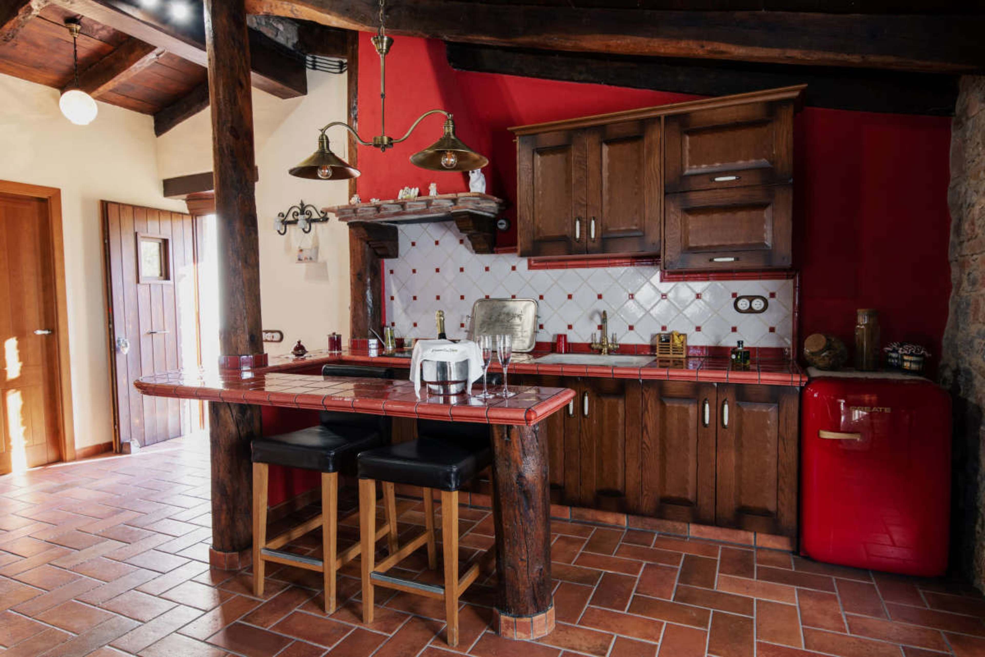 A rustic kitchen features wood cabinets, a red retro refrigerator, and a bar with two stools under hanging lights.