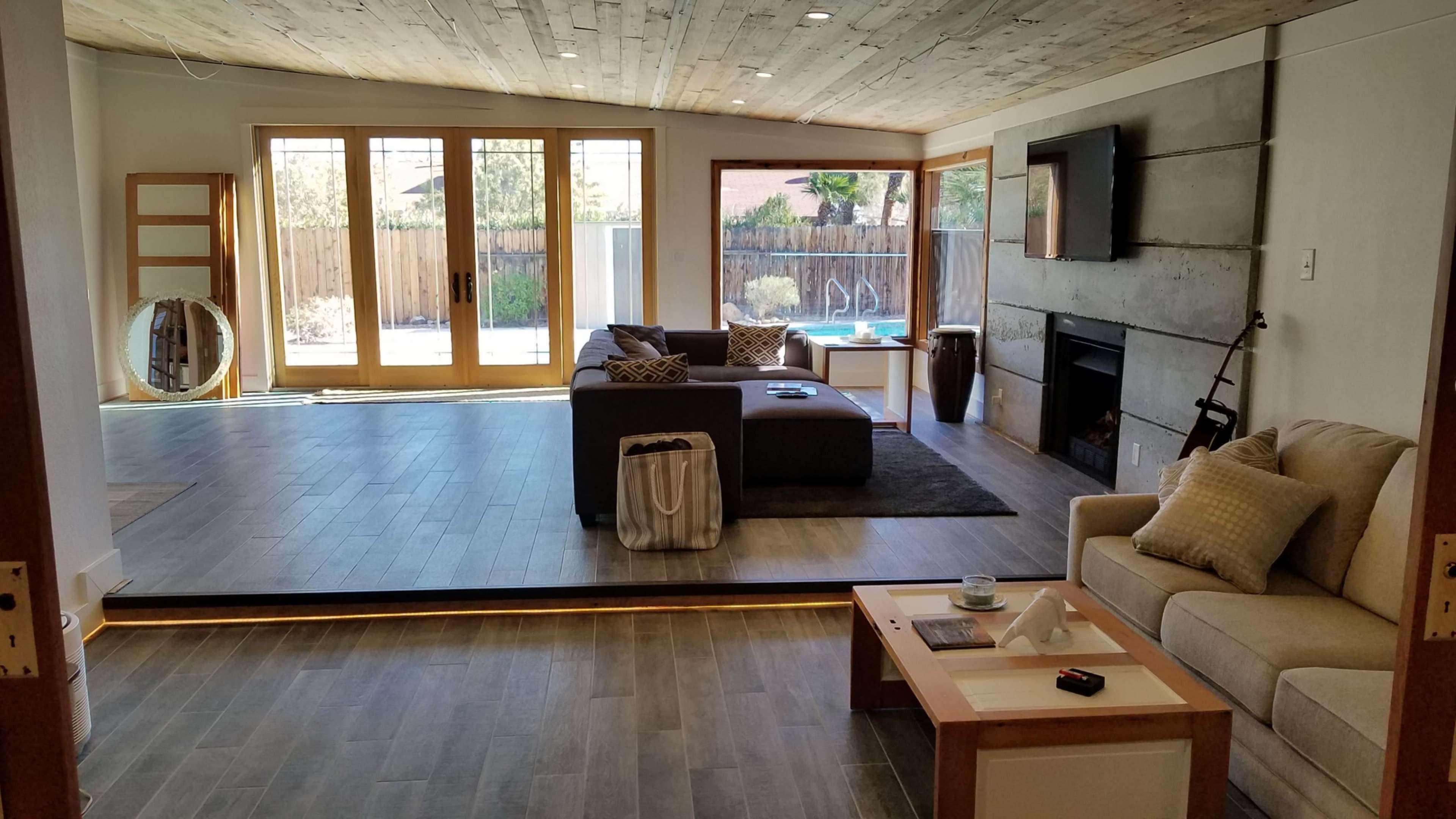 The image shows a modern living room with a sectional sofa, a coffee table, and large windows overlooking a backyard with a pool.