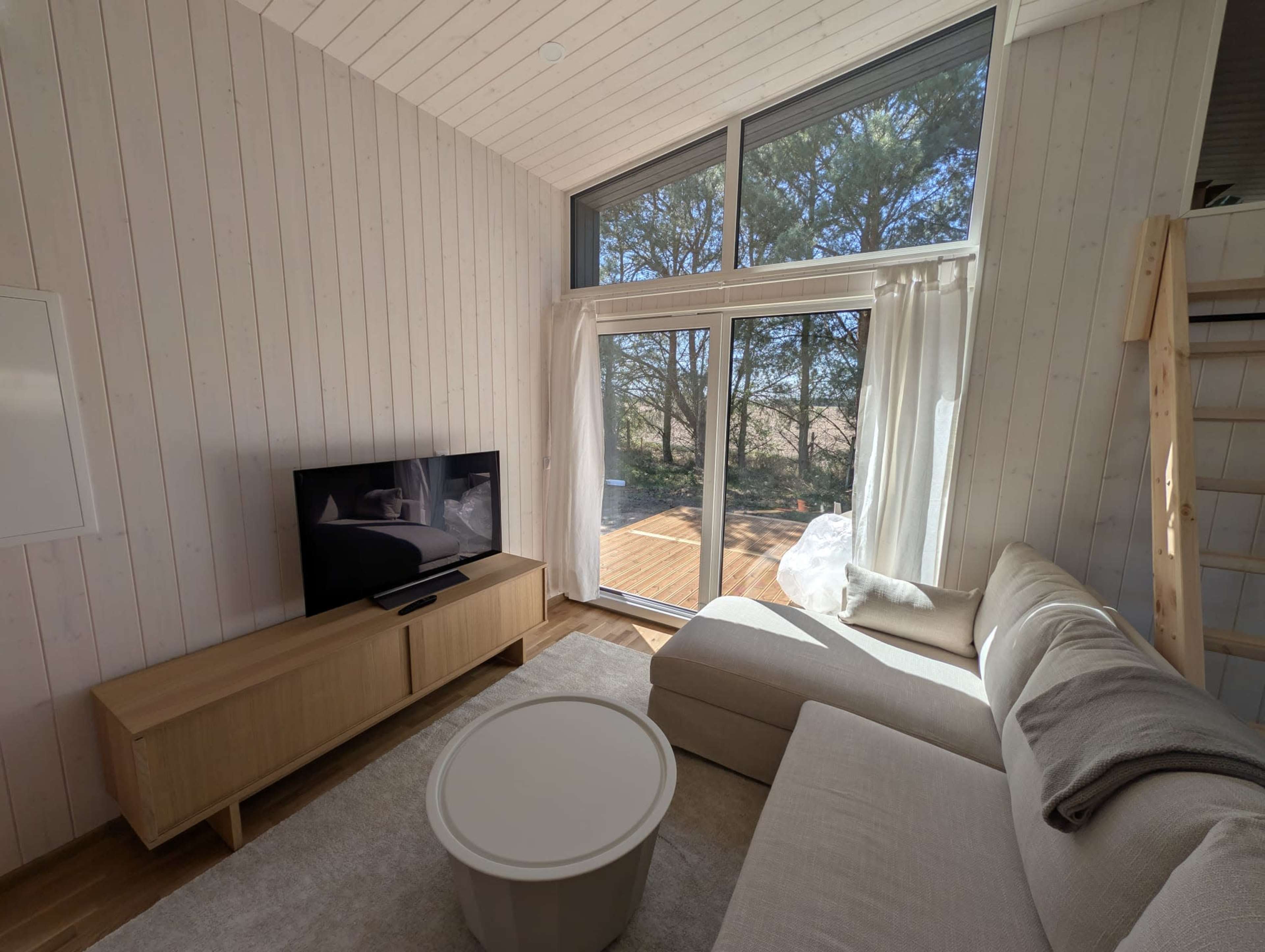 The image shows a modern living room with a sectional sofa, coffee table, and large windows overlooking a wooden deck and trees outside.