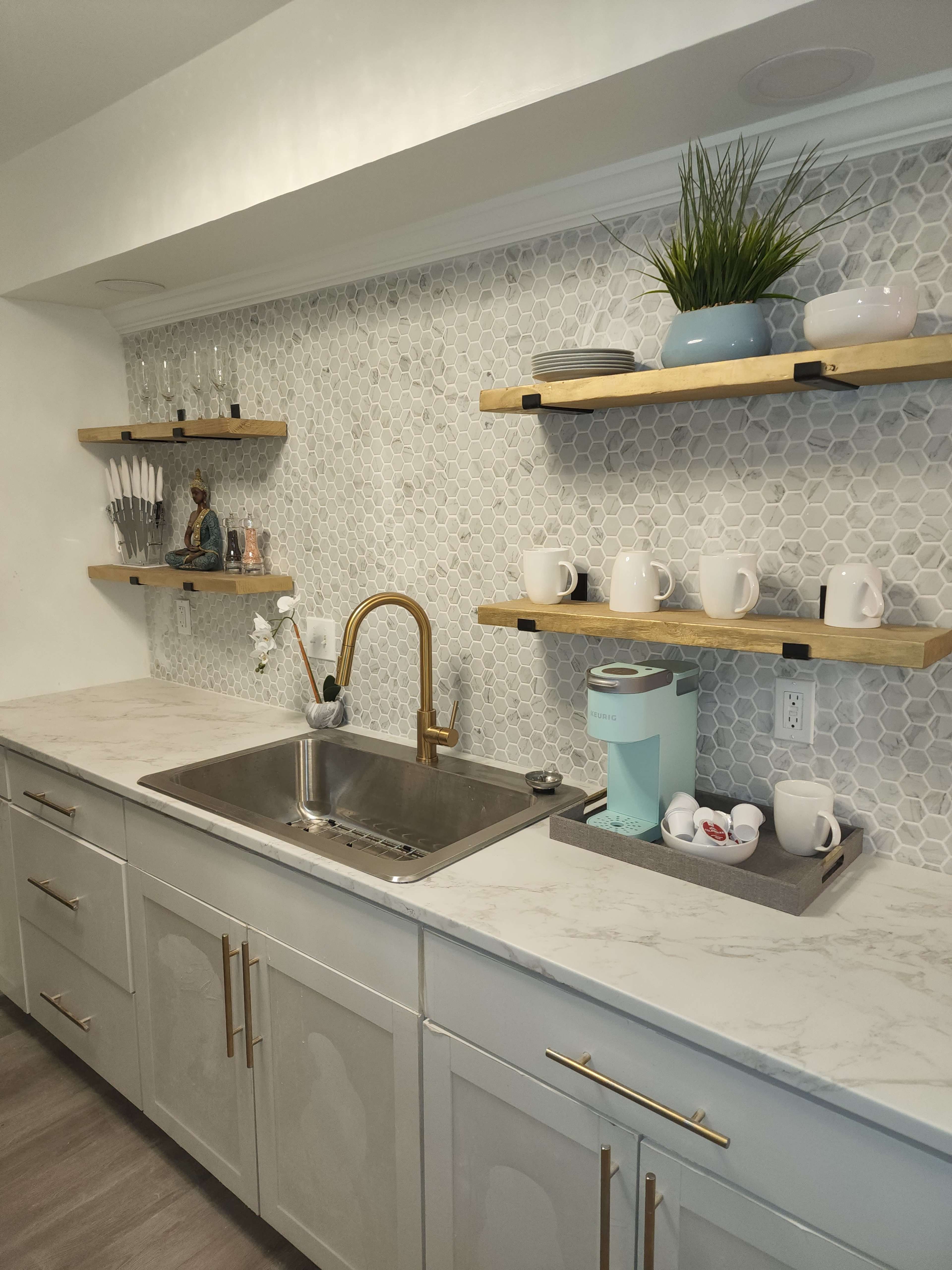 A modern kitchen featuring a stainless steel sink, light gray cabinets, hexagonal tiled backsplash, and wooden open shelves displaying dishes and utensils.