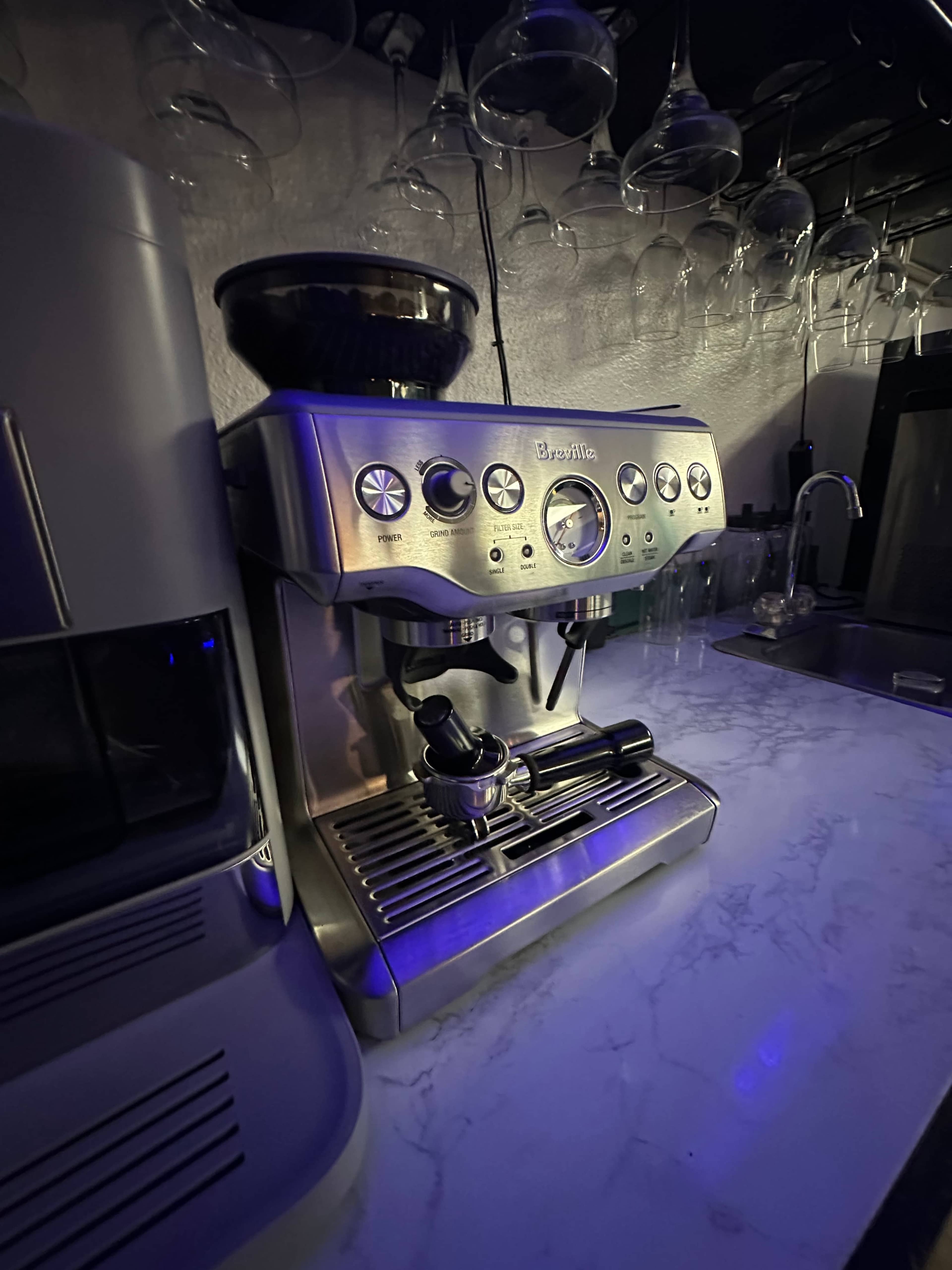 A stainless steel espresso machine is positioned on a marble countertop, with wine glasses hanging above it in a dimly lit kitchen.