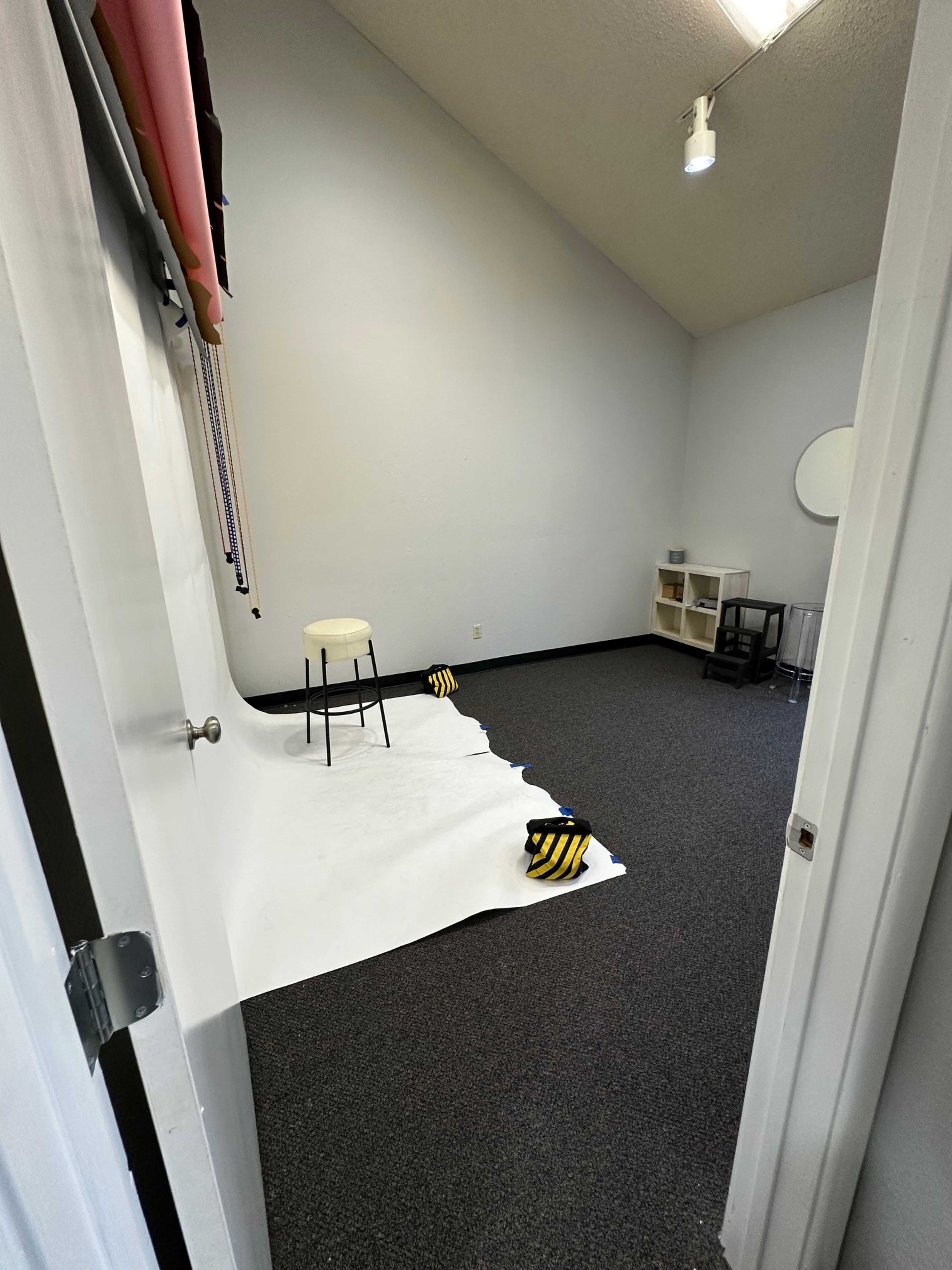 The image shows an empty photography studio with a white backdrop, a stool, and black and yellow striped props on the floor.