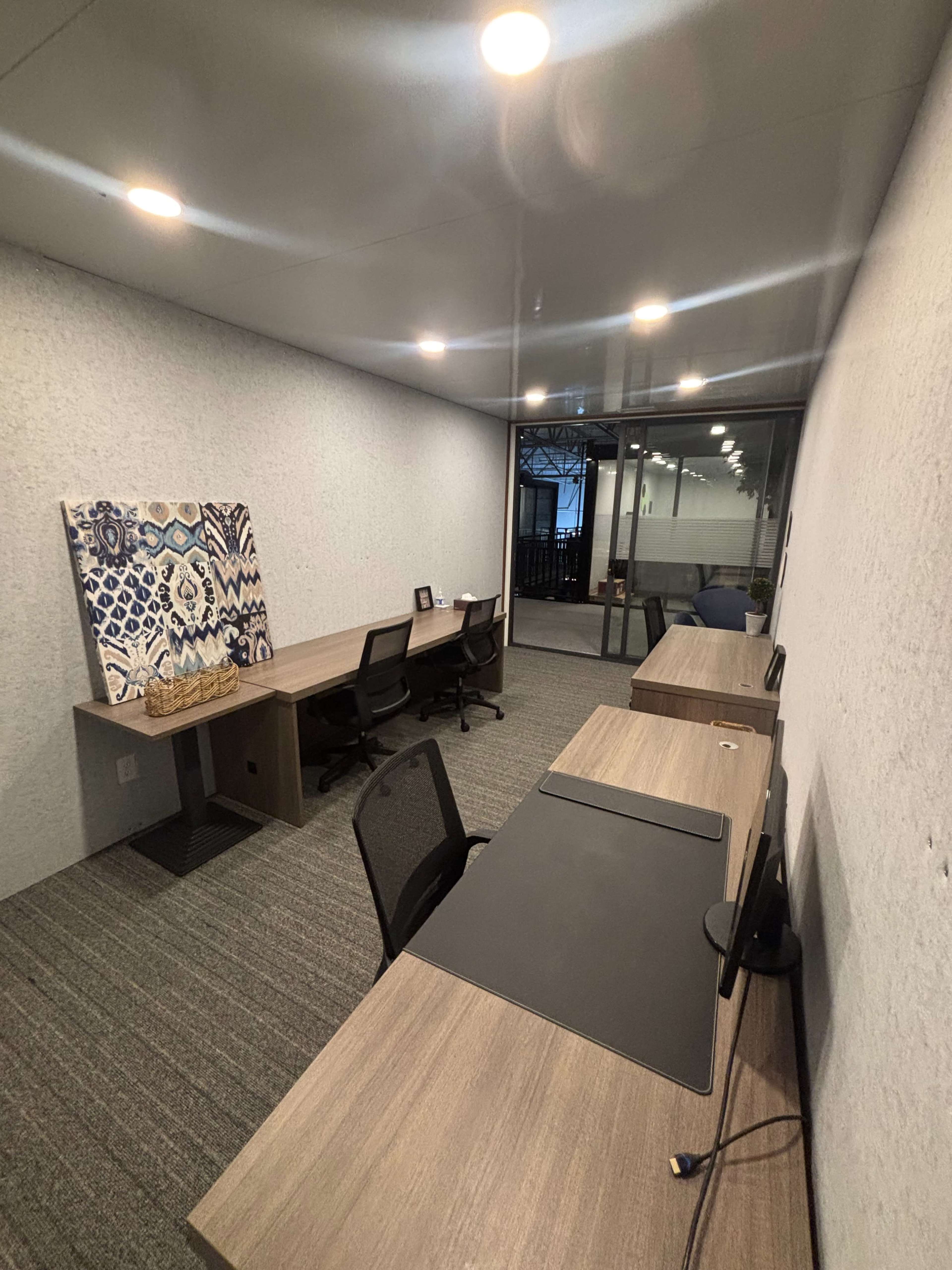 The image shows a modern office space with two workstations, a piece of art on the wall, and large windows allowing natural light.