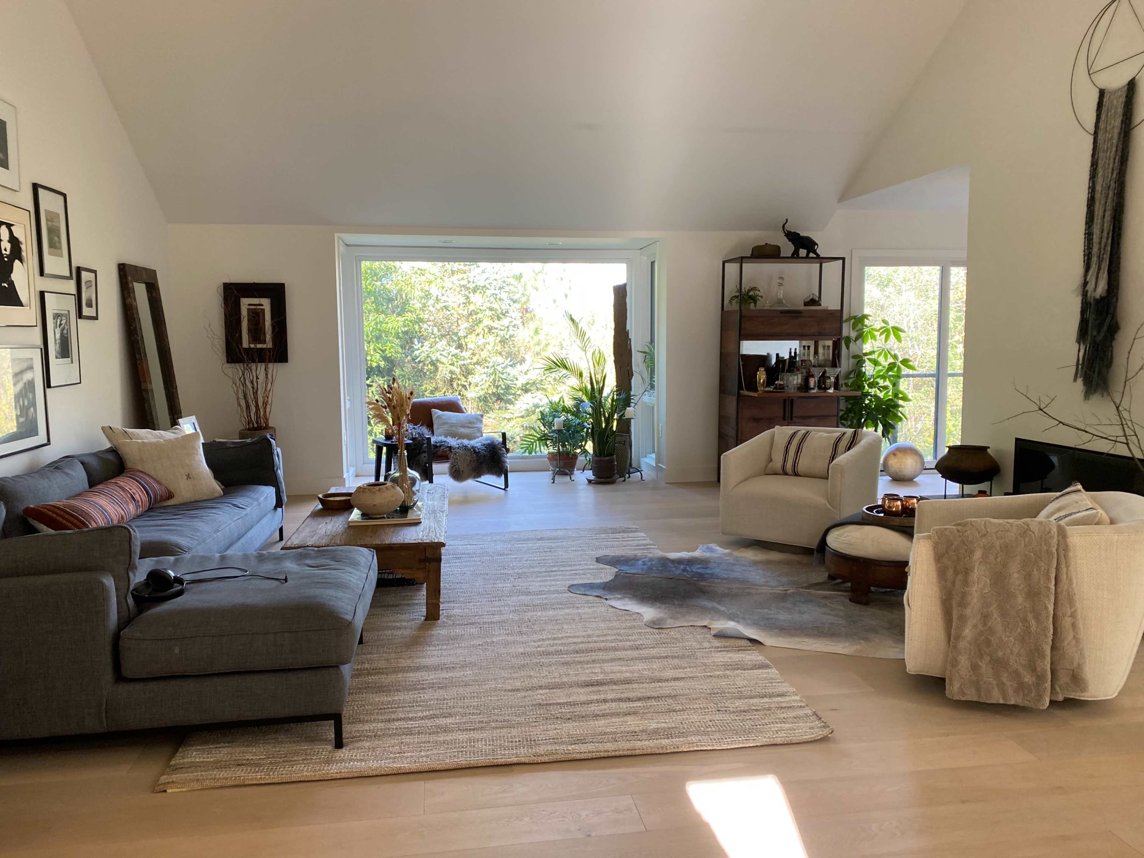 A modern living room with a large window overlooking greenery, featuring a gray sofa, a round coffee table, and a textured area rug on a light wood floor.