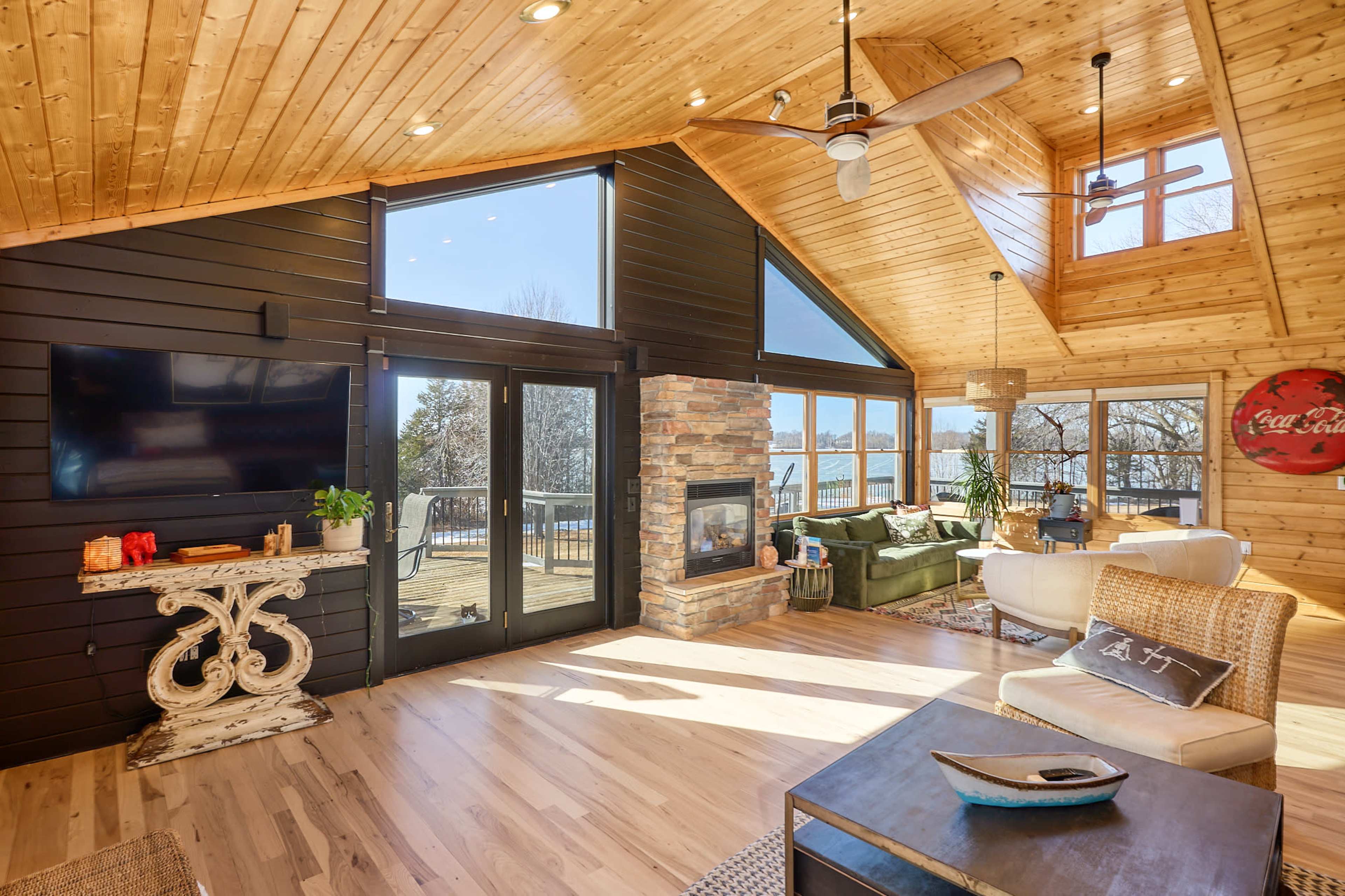 The image shows a cozy, modern living room with wooden ceilings, large windows overlooking a deck, and a stone fireplace.