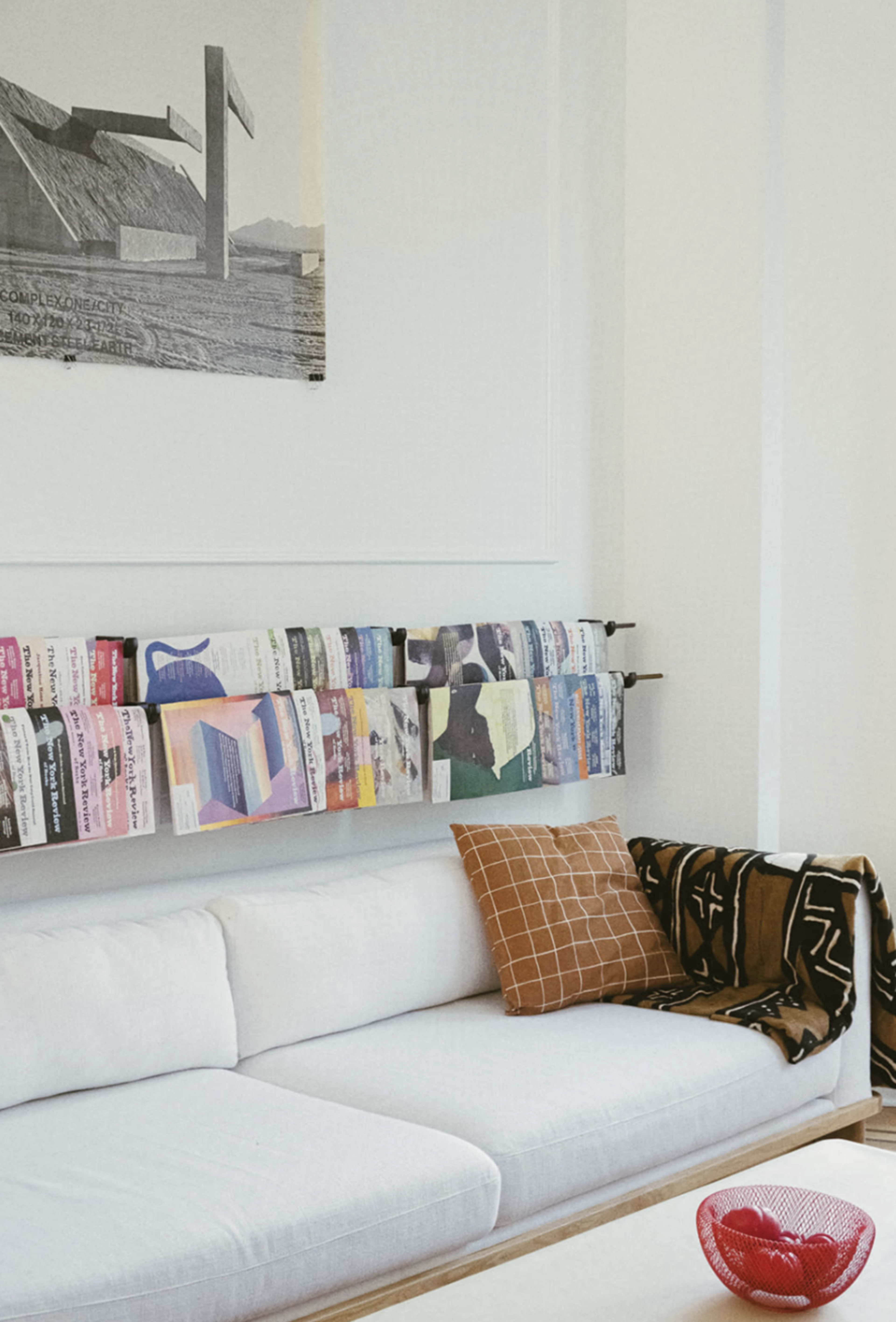The image shows a modern living room with a white sofa, a colorful magazine display on the wall, and a red bowl on a wooden table.