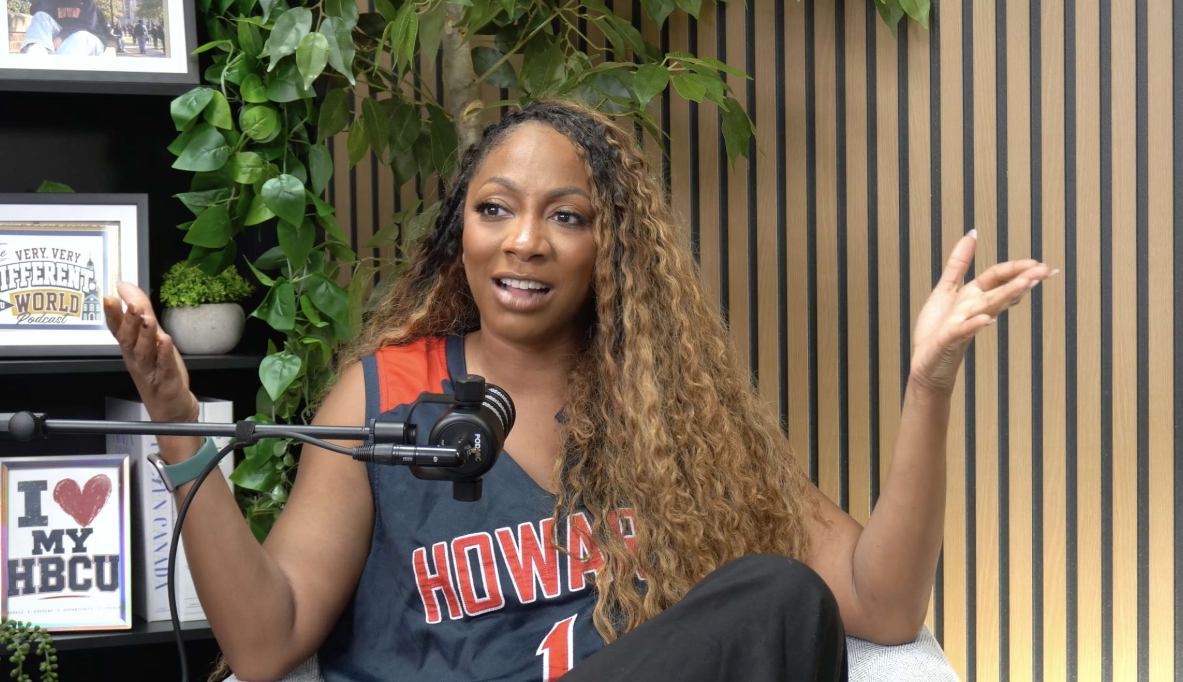 A woman with long, curly hair wearing a Howard University jersey is speaking animatedly in a studio setting with indoor plants in the background.
