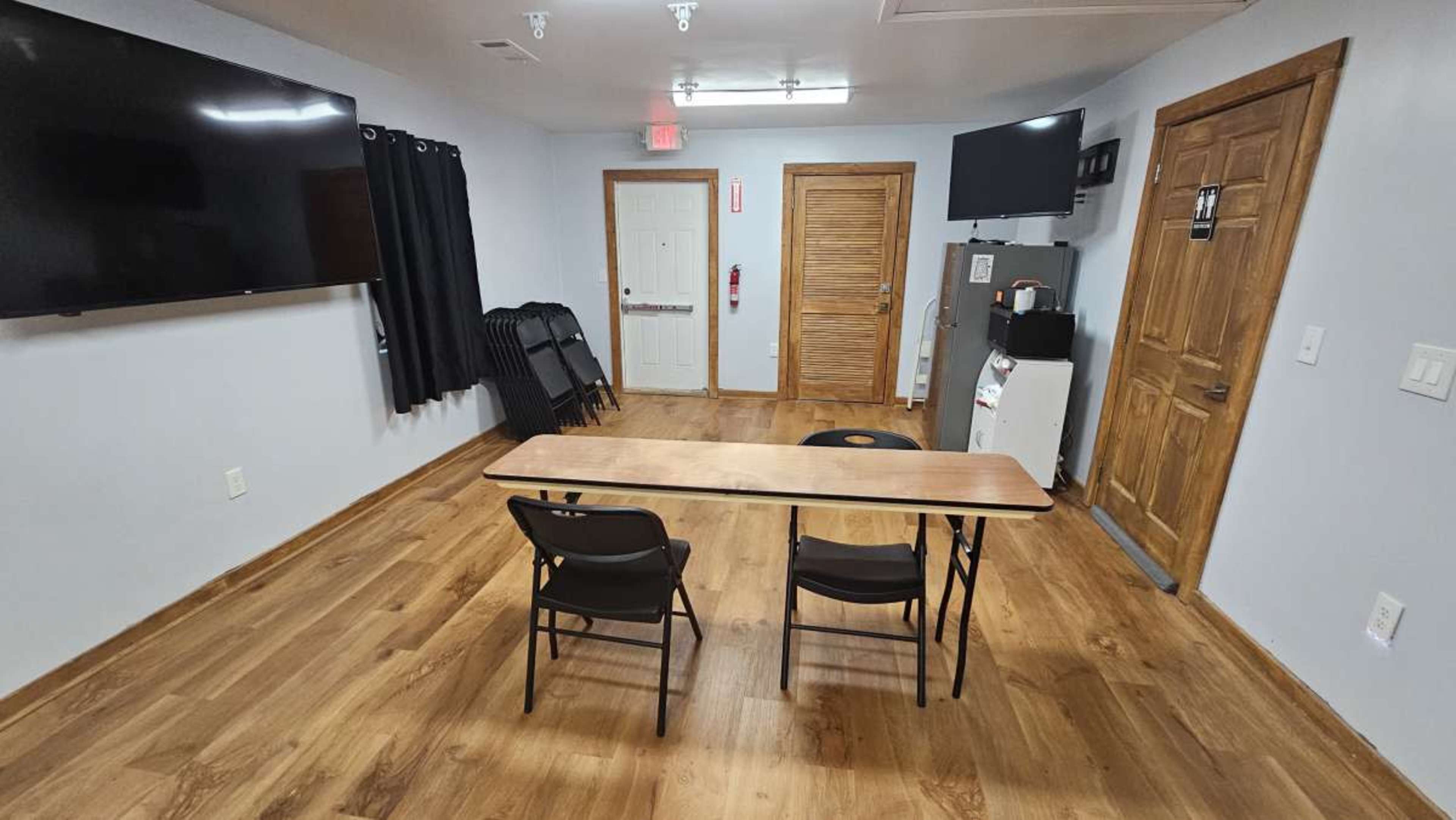 The image displays a meeting room with a wooden table and two chairs, walls painted light blue, a large TV, and a small fridge in the corner.
