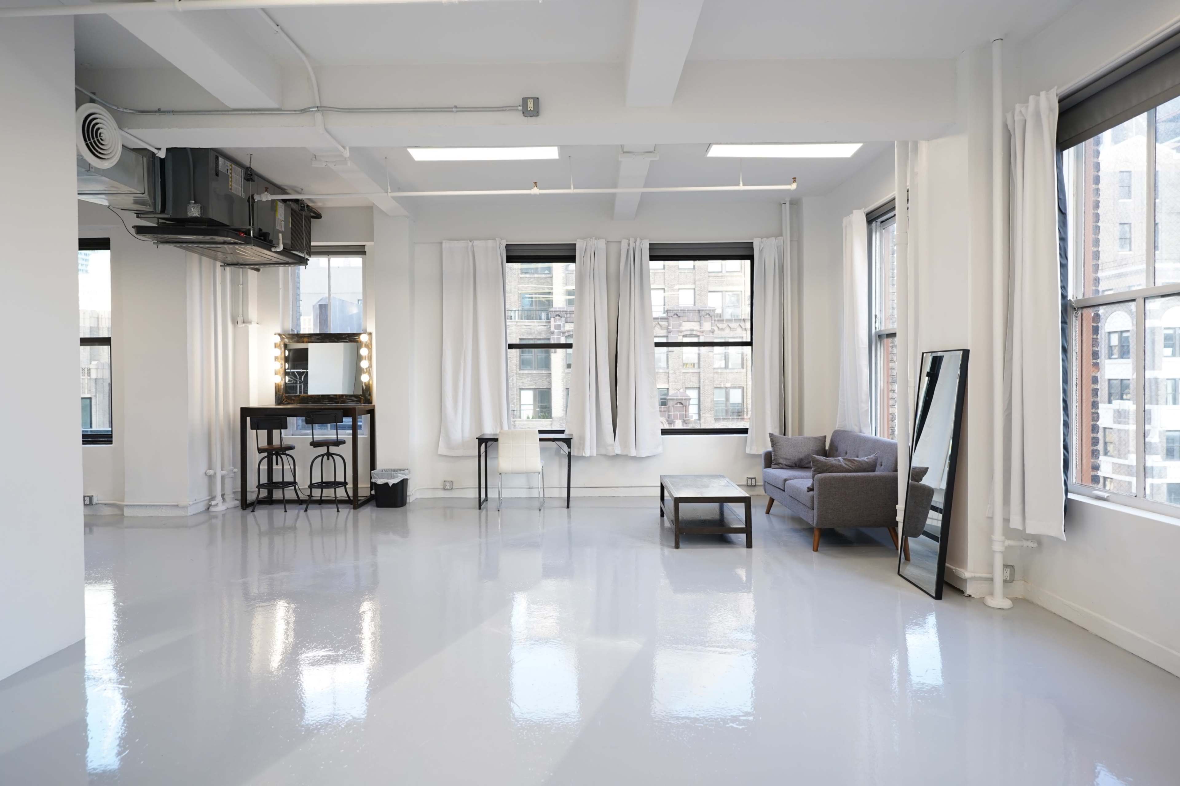 The image shows a bright, spacious room with large windows, a mirror, a table and chairs, and a sofa, all set against a clean, polished floor.