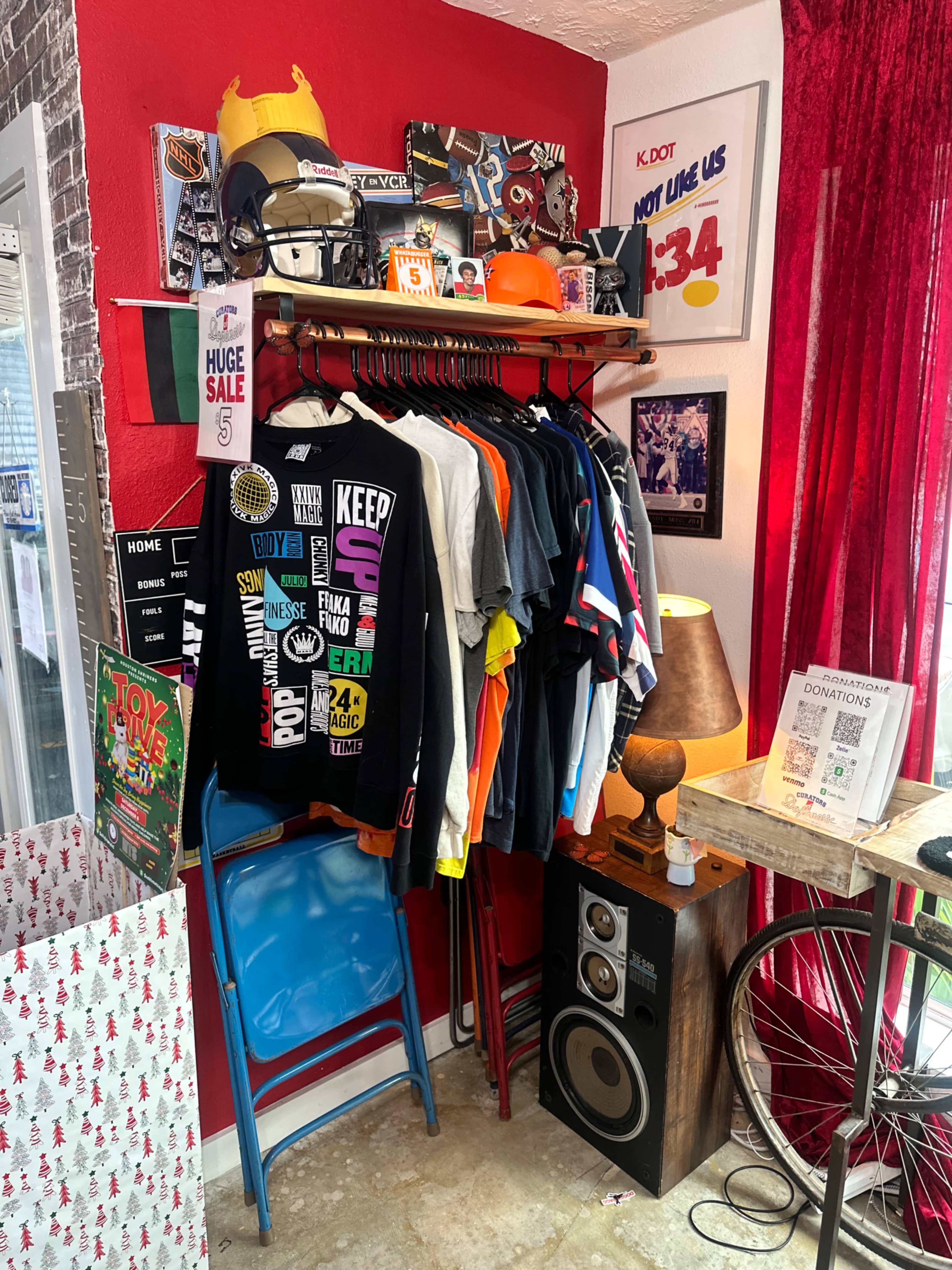 The image shows a clothing display rack filled with various T-shirts, alongside a blue folding chair and a lamp on a wooden stand, with a bicycle in the background.