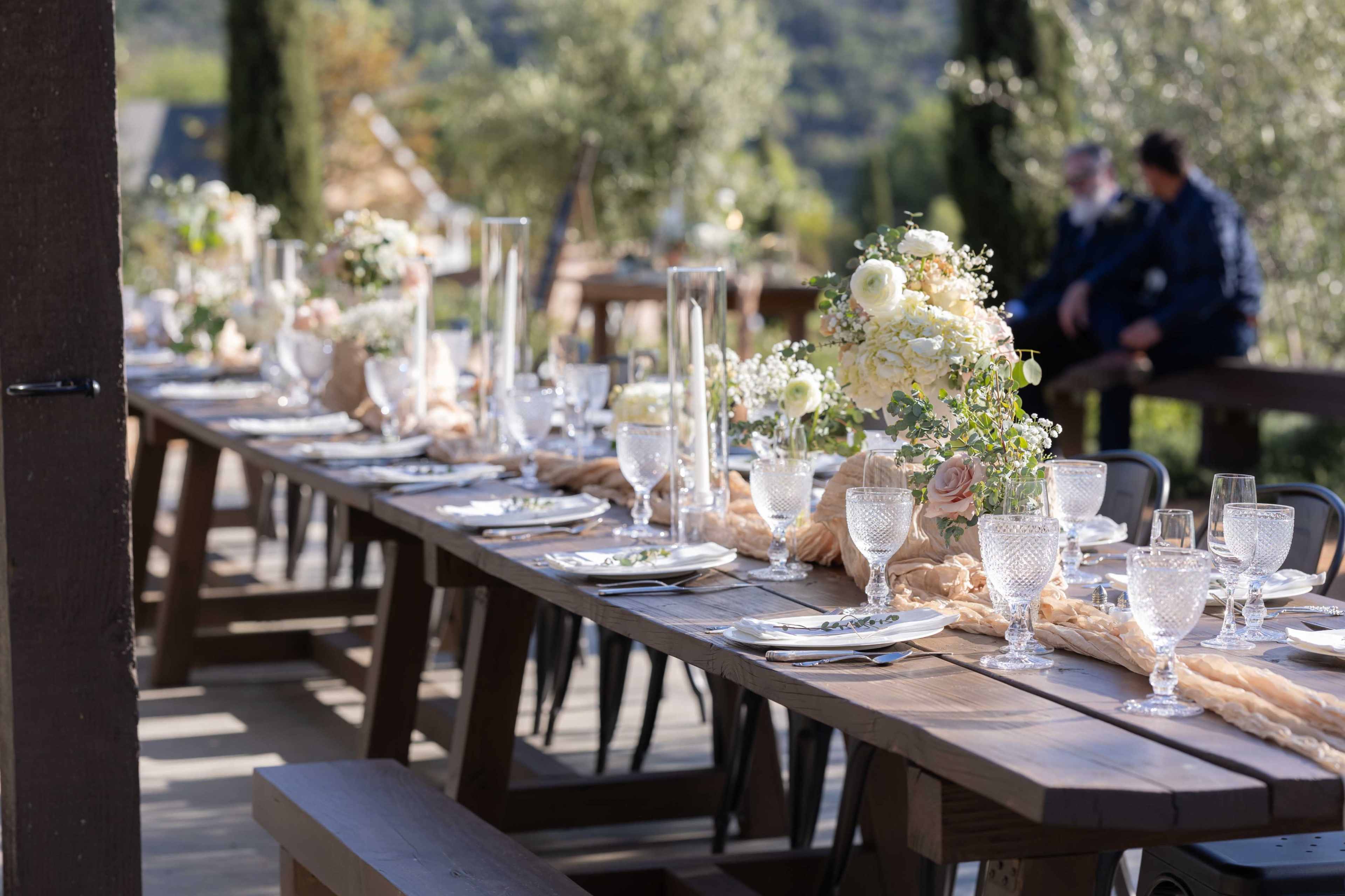 The image features a long wooden table set with elegant glassware and floral centerpieces, surrounded by a scenic outdoor venue.