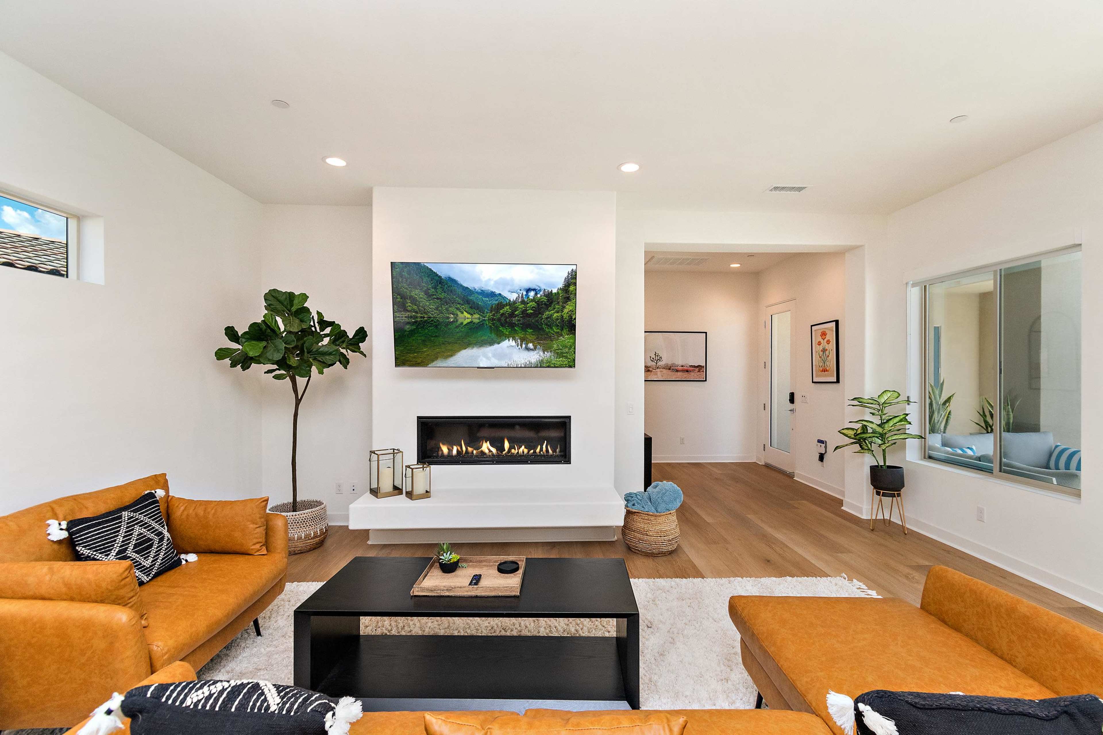 The living room features two orange leather sofas facing a dark coffee table, with a mounted television above a modern fireplace and large windows with greenery.