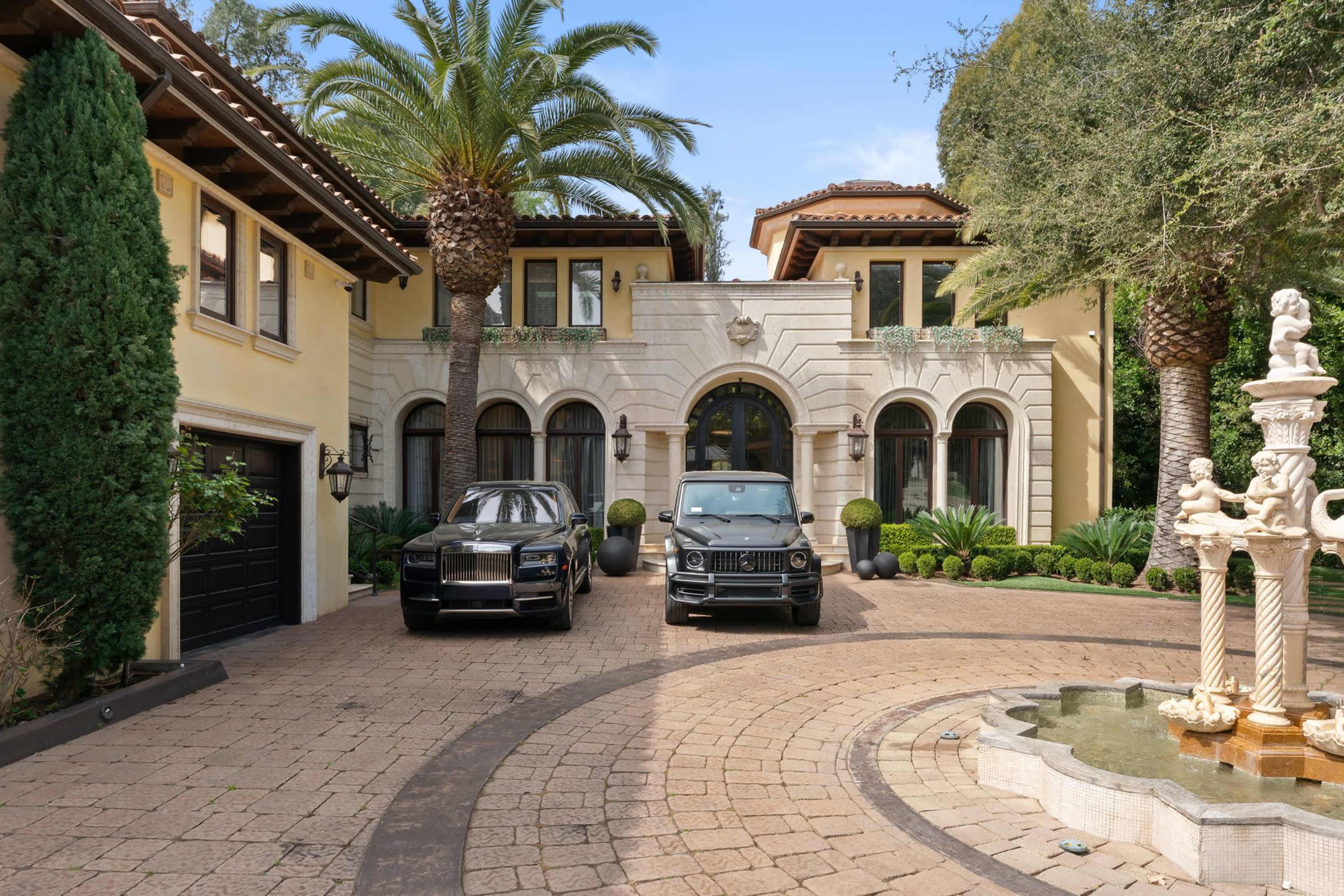 The entrance of a luxurious house features two high-end vehicles parked in a circular driveway surrounded by palm trees and manicured landscaping.