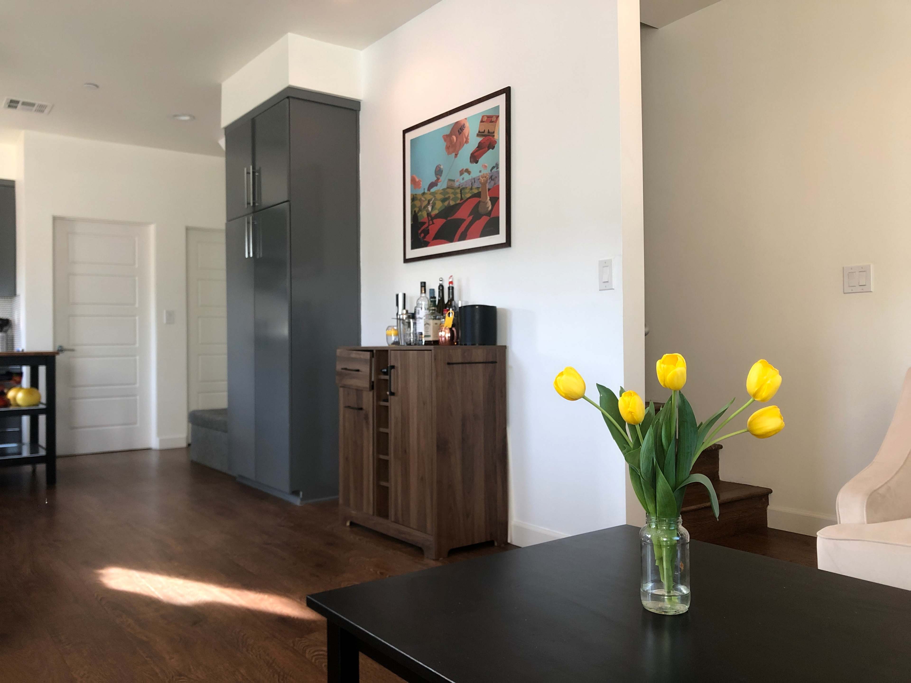 A vase of yellow tulips sits on a black table in a bright living space with a bar cabinet and stairs visible in the background.