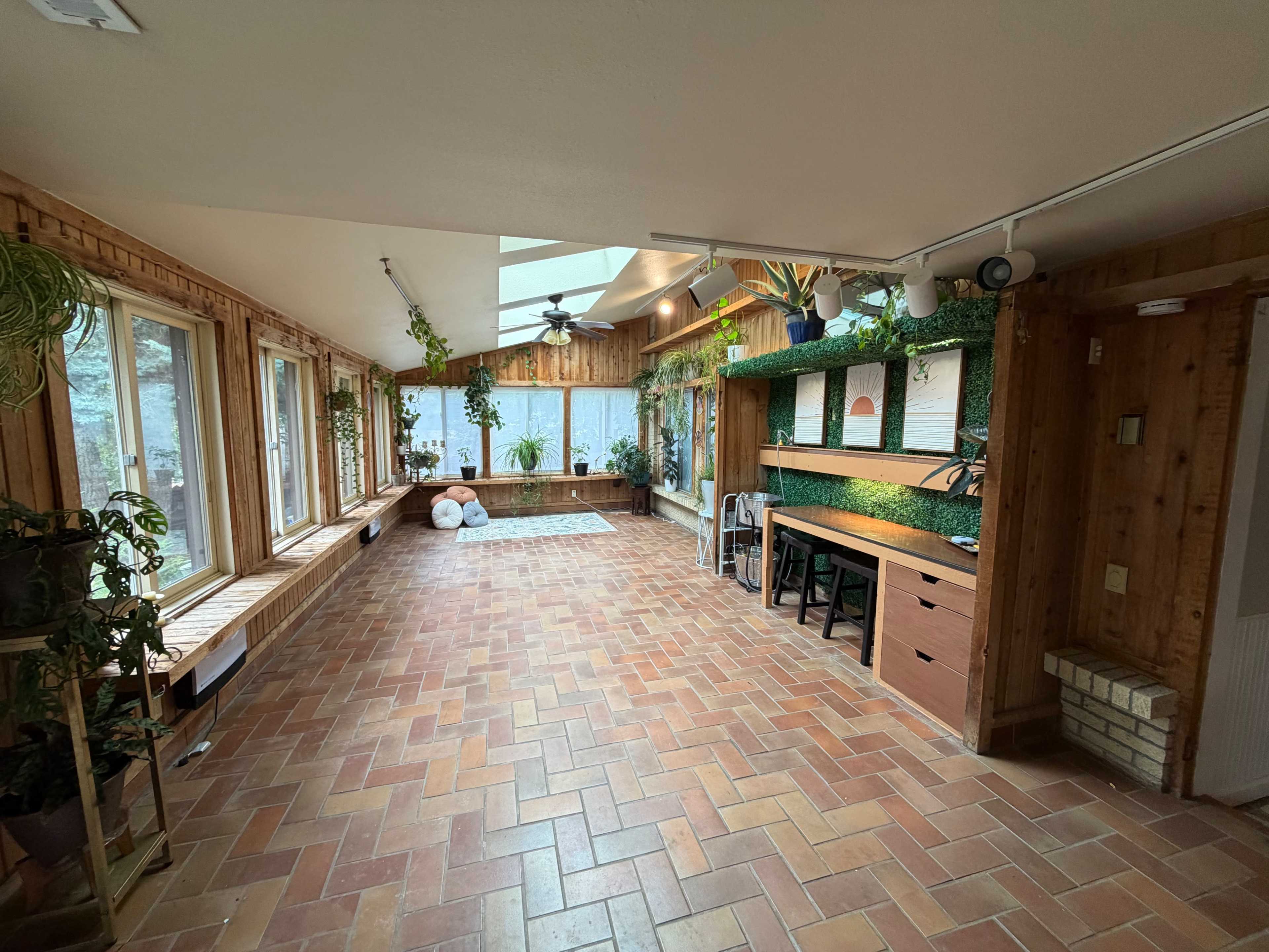 The image shows a spacious sunroom with terracotta tile flooring, large windows, and a wooden interior, featuring plants arranged on shelves and a work area with a desk.