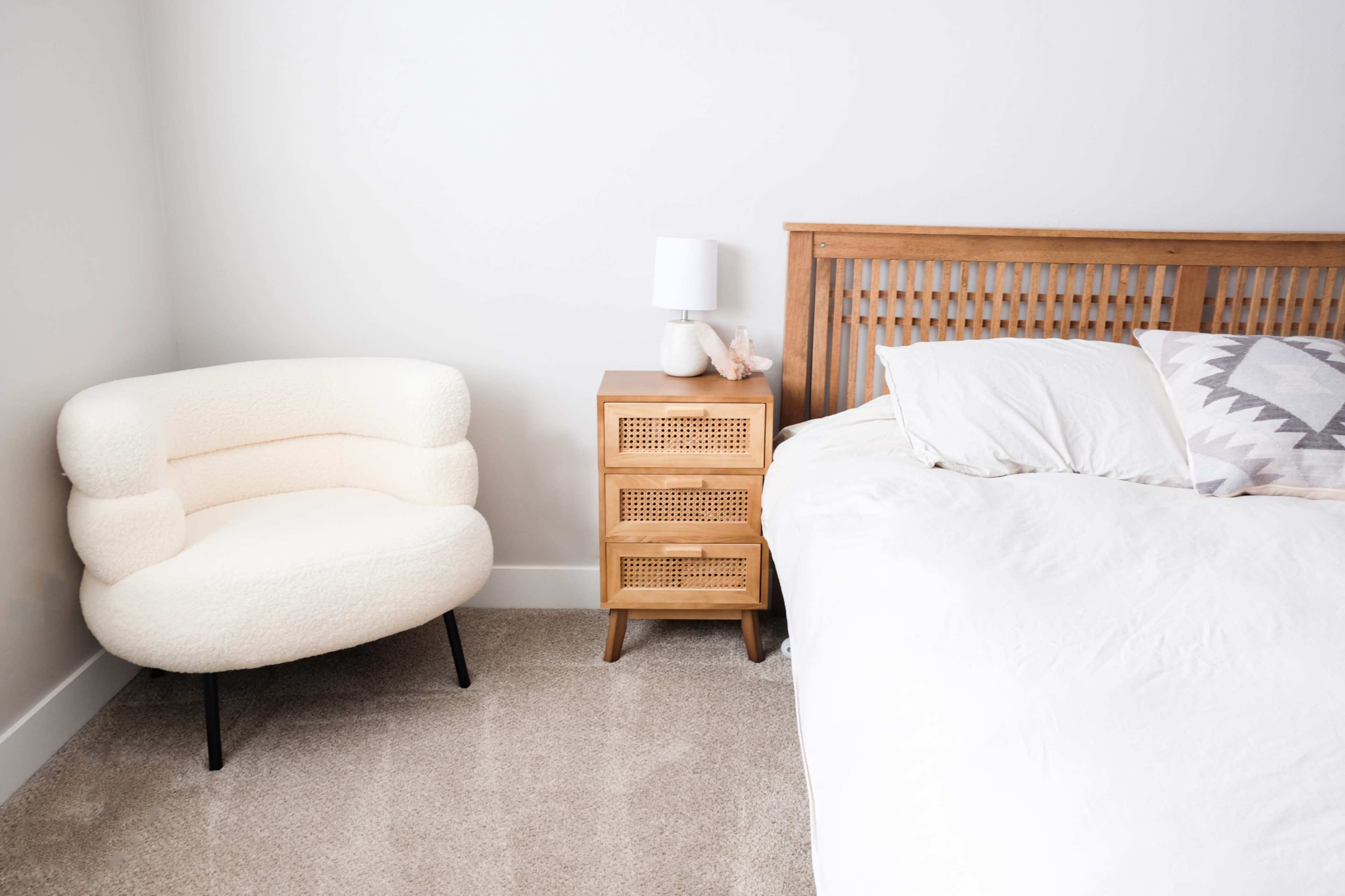 A light-colored bedroom features a wooden bed, a bedside table with three drawers, and a plush, cream-colored chair in the corner.