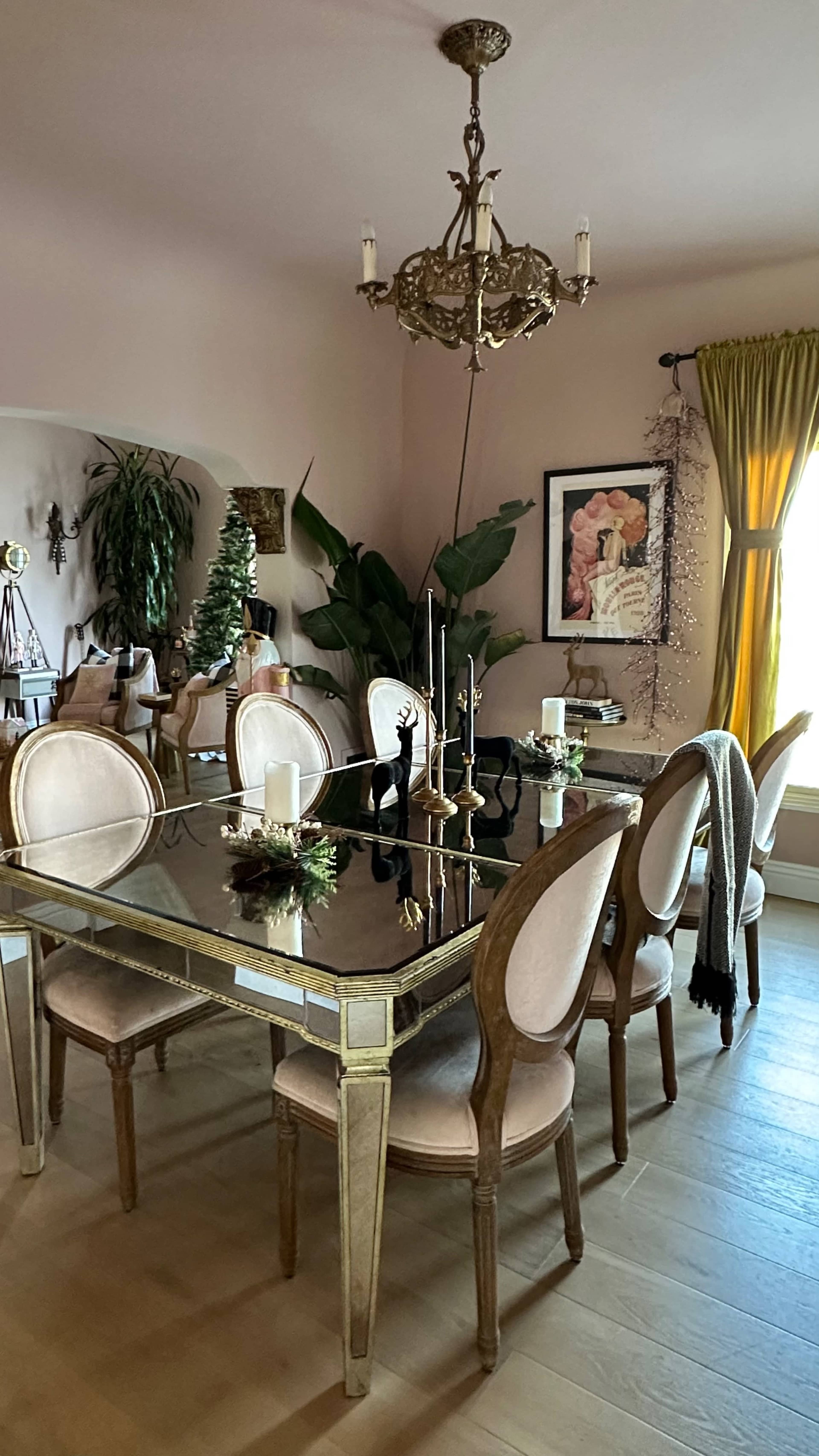 A dining room features a large black table surrounded by upholstered chairs, with a chandelier overhead and plants in the corners.