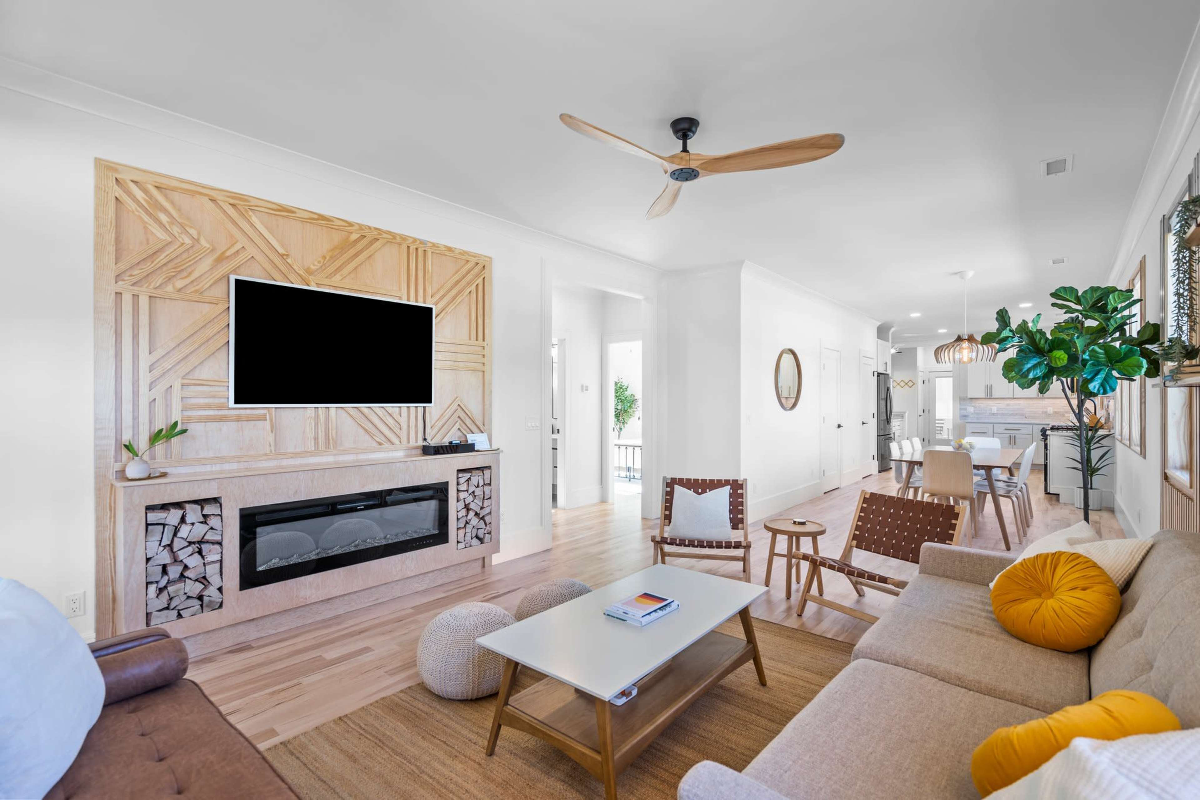 A modern living room features a wall-mounted TV above a decorative fireplace, surrounded by light-colored furniture and plants.