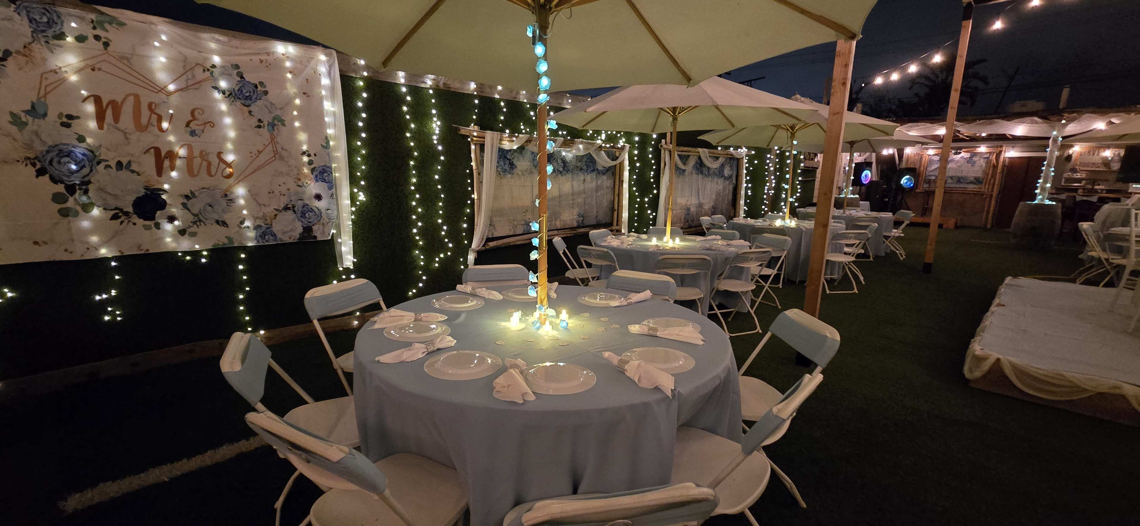 The image shows a decorated outdoor event space with tables covered in light blue tablecloths, white chairs, and string lights illuminating the area.