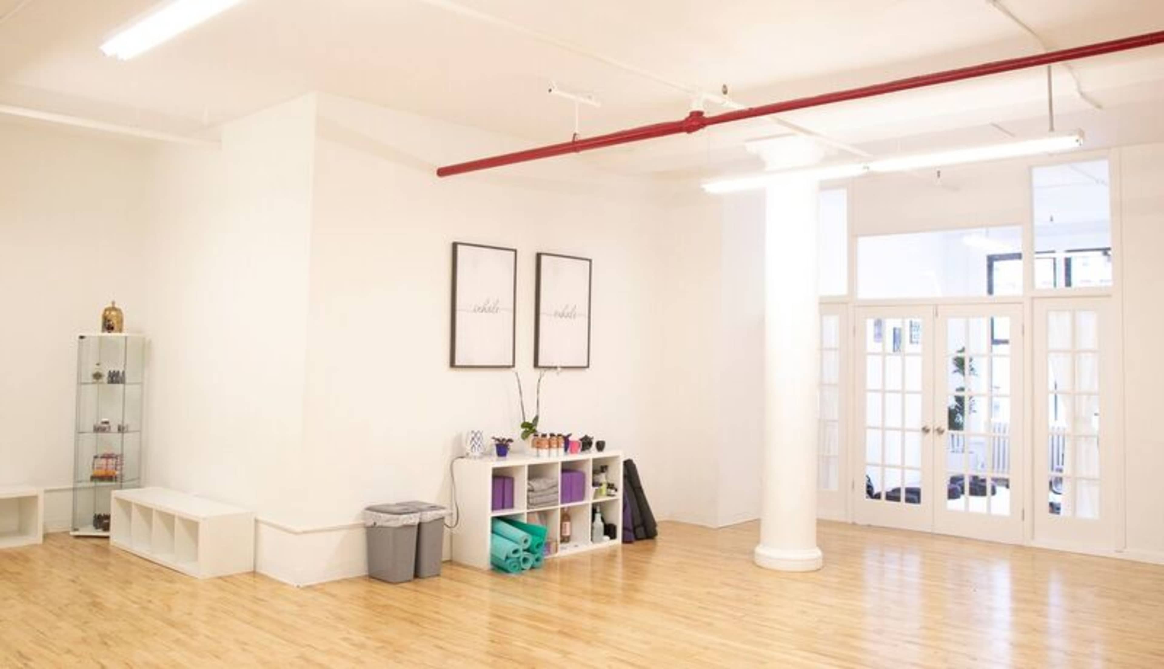 A spacious, uncluttered room with light-colored walls, wooden flooring, and large windows, featuring shelves for storage and exercise mats.