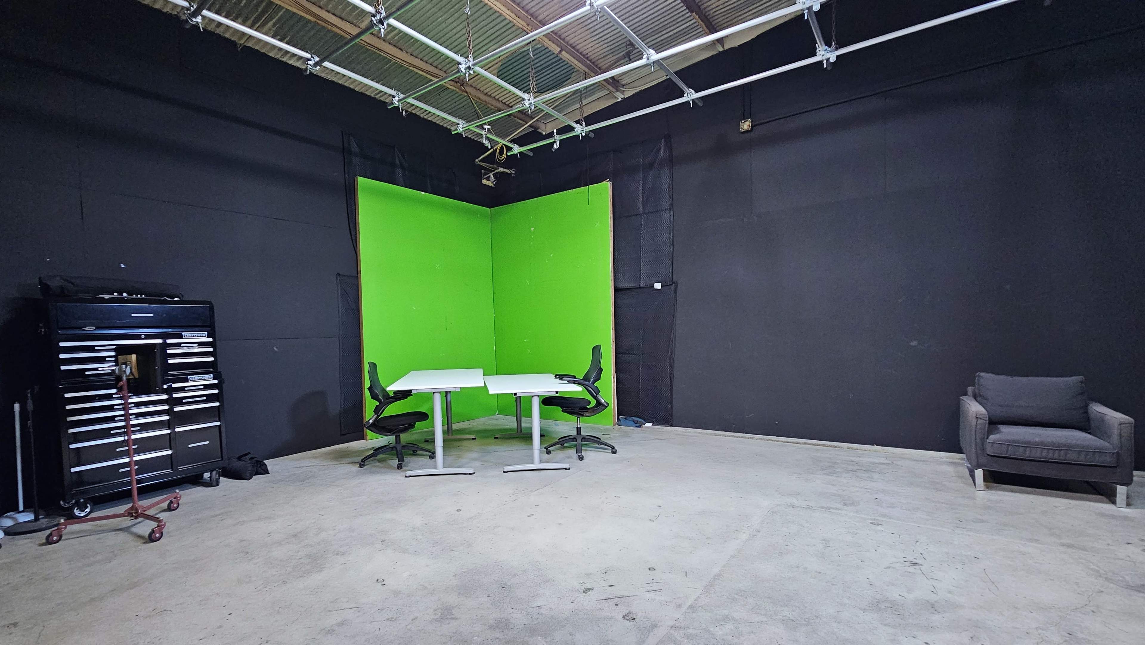 A studio space with a green screen backdrop, a pair of white tables with black chairs, a gray armchair, and a tool storage unit against the black walls.