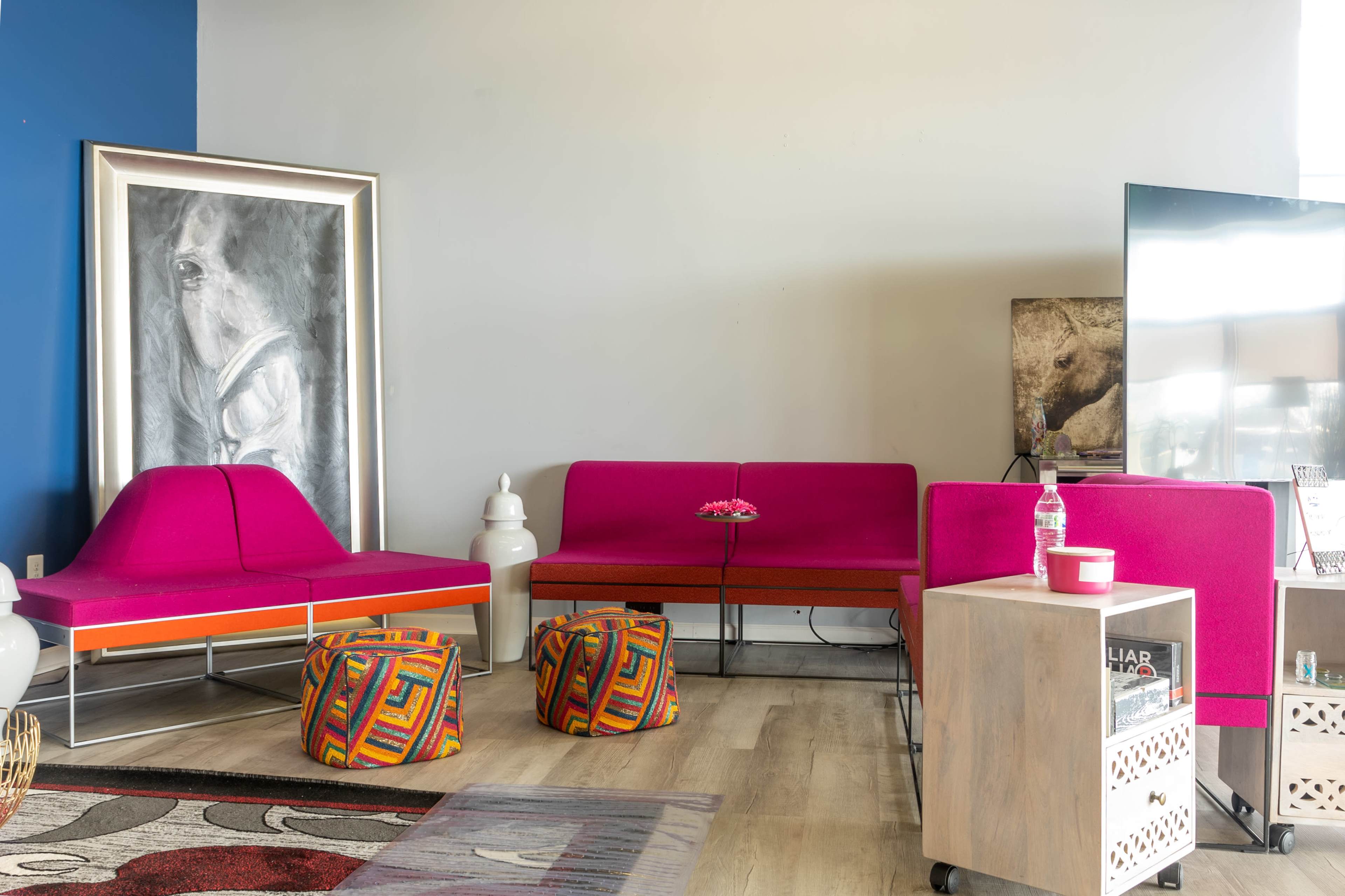 The scene features a modern living area with pink upholstered seating, colorful patterned poufs, a large framed artwork, and a television on a side cart.