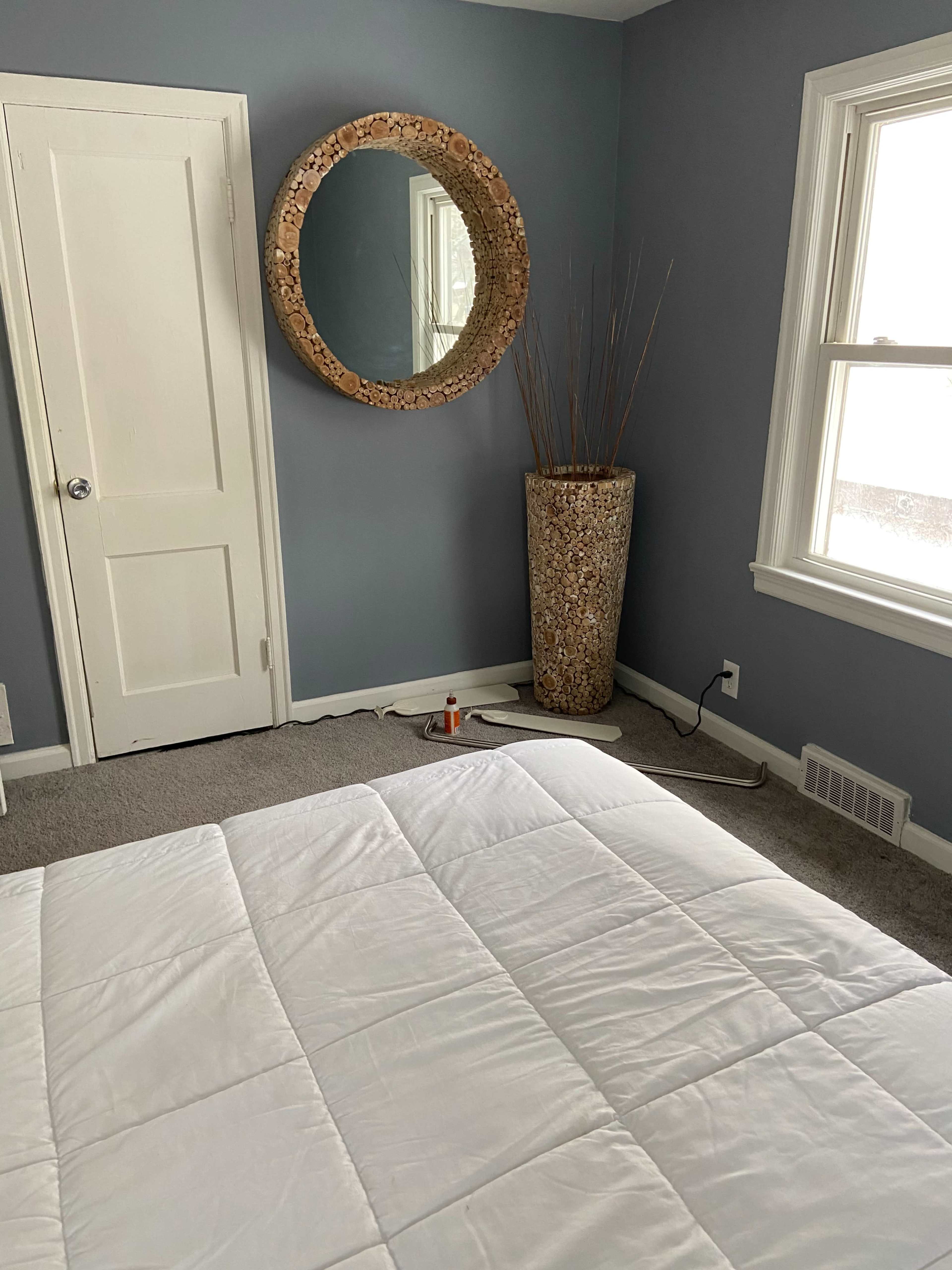 A simple bedroom with a bed covered by a white quilt, a circular mirror on the wall, and a large decorative vase in the corner near a closed door.