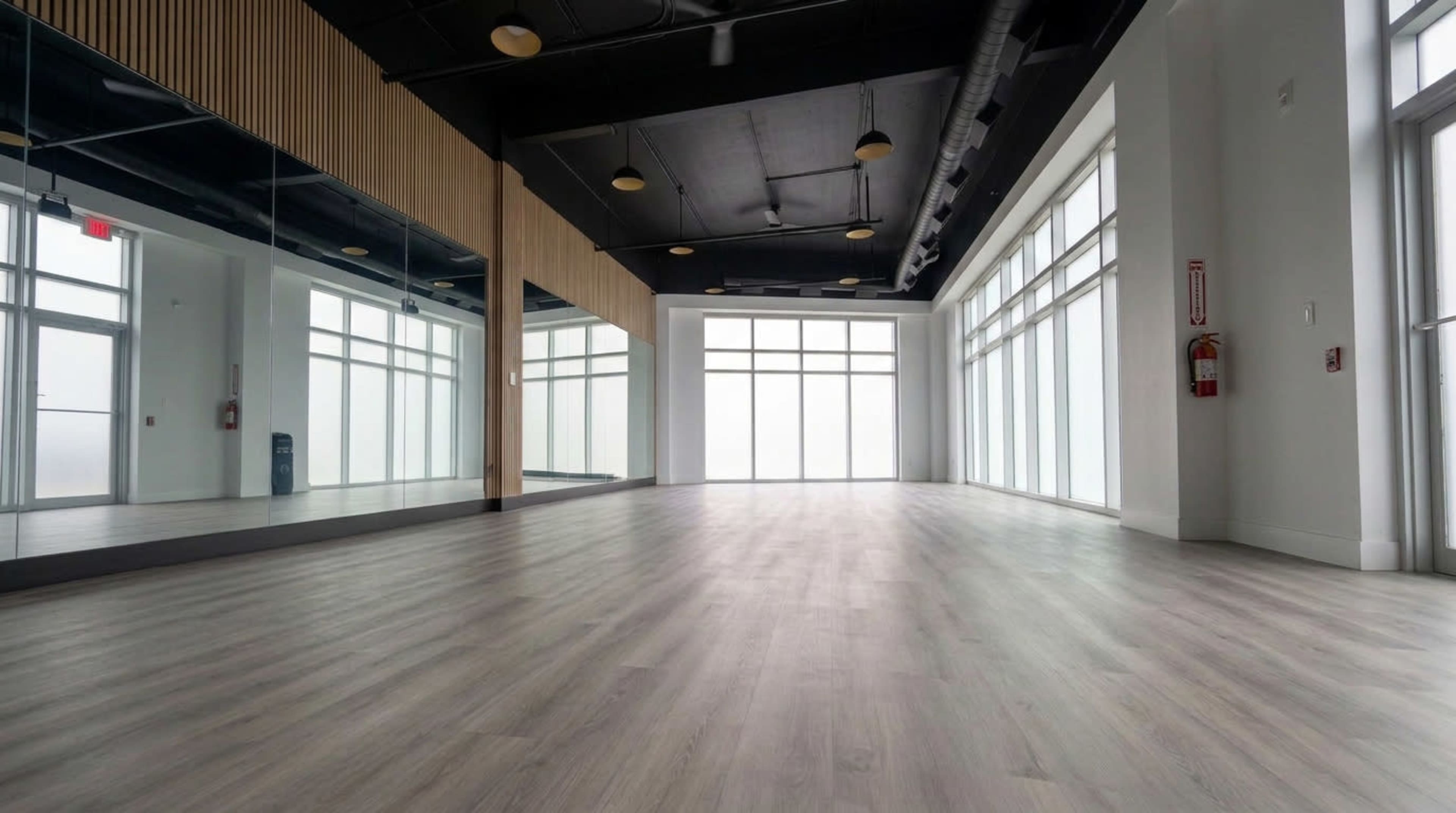 The image shows a spacious, empty room with large windows, wooden accents, and a polished floor.