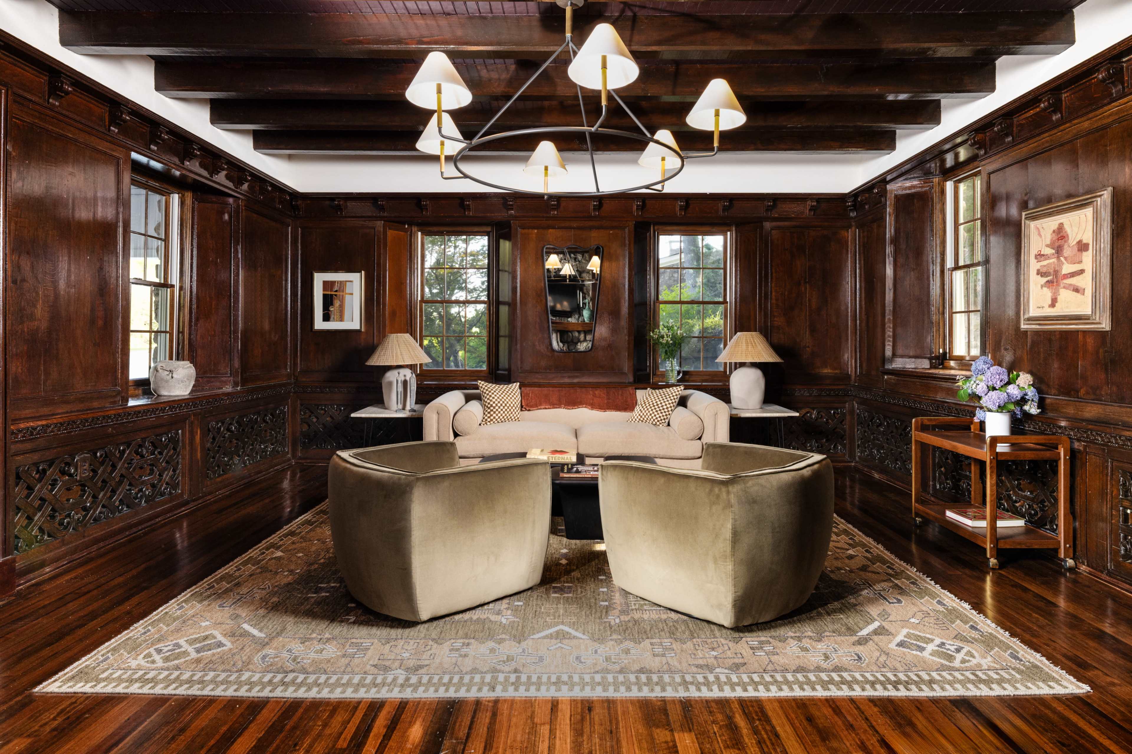 A cozy living room with dark wooden paneling, a central chandelier, two green armchairs facing a beige sofa, and a decorative area rug.