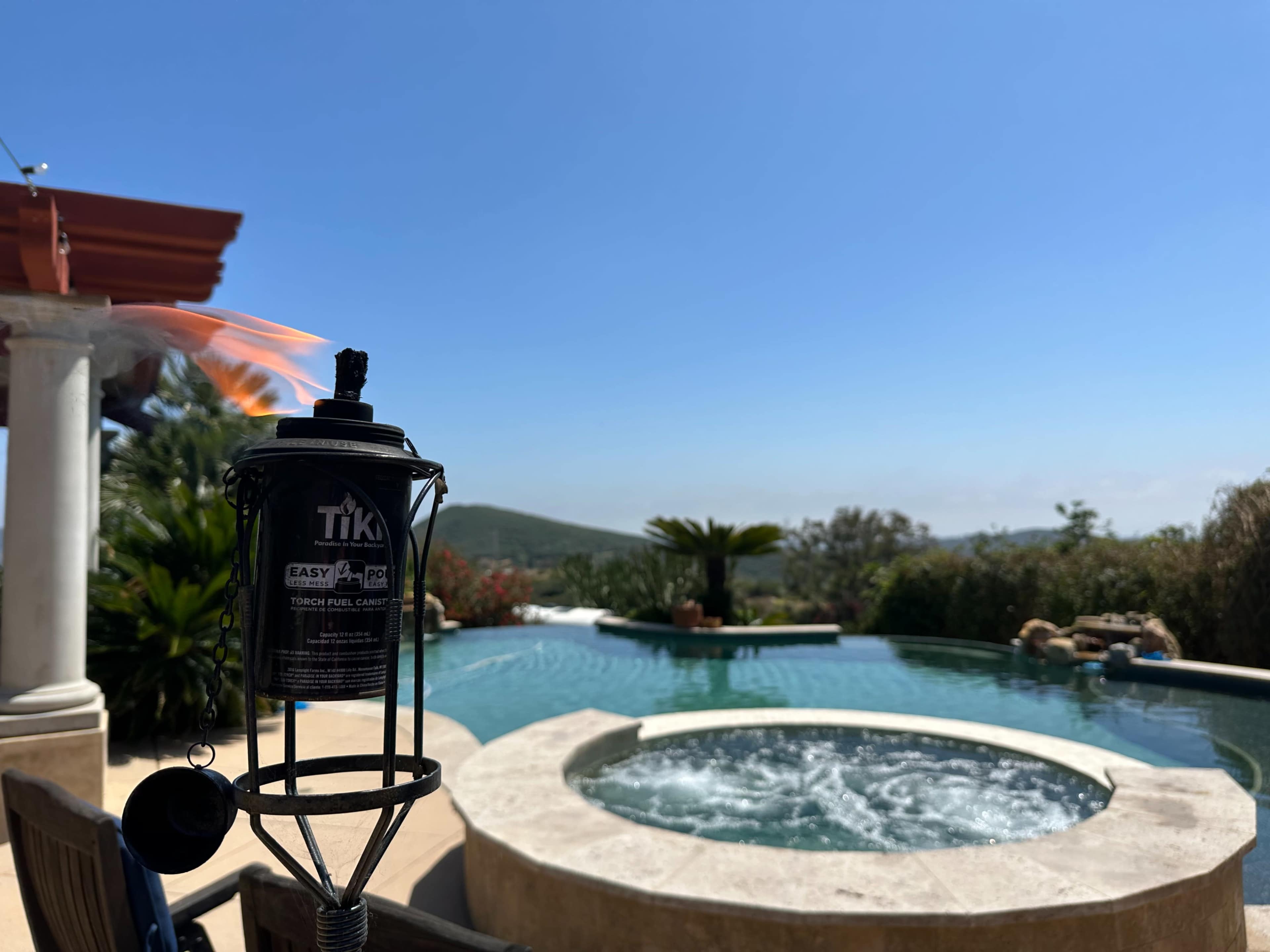 A tiki torch burns brightly near a pool and hot tub surrounded by lush greenery and hills under a clear blue sky.