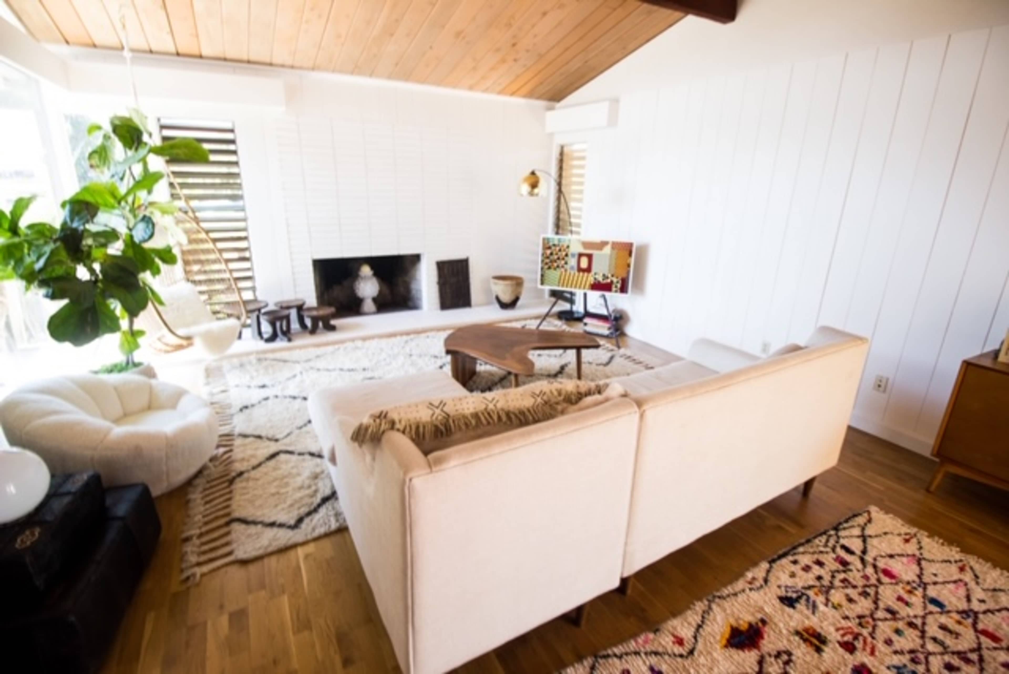 The image shows a minimalist living room featuring a light-colored sofa, a wooden coffee table, a large area rug, and a potted plant beside a fireplace.