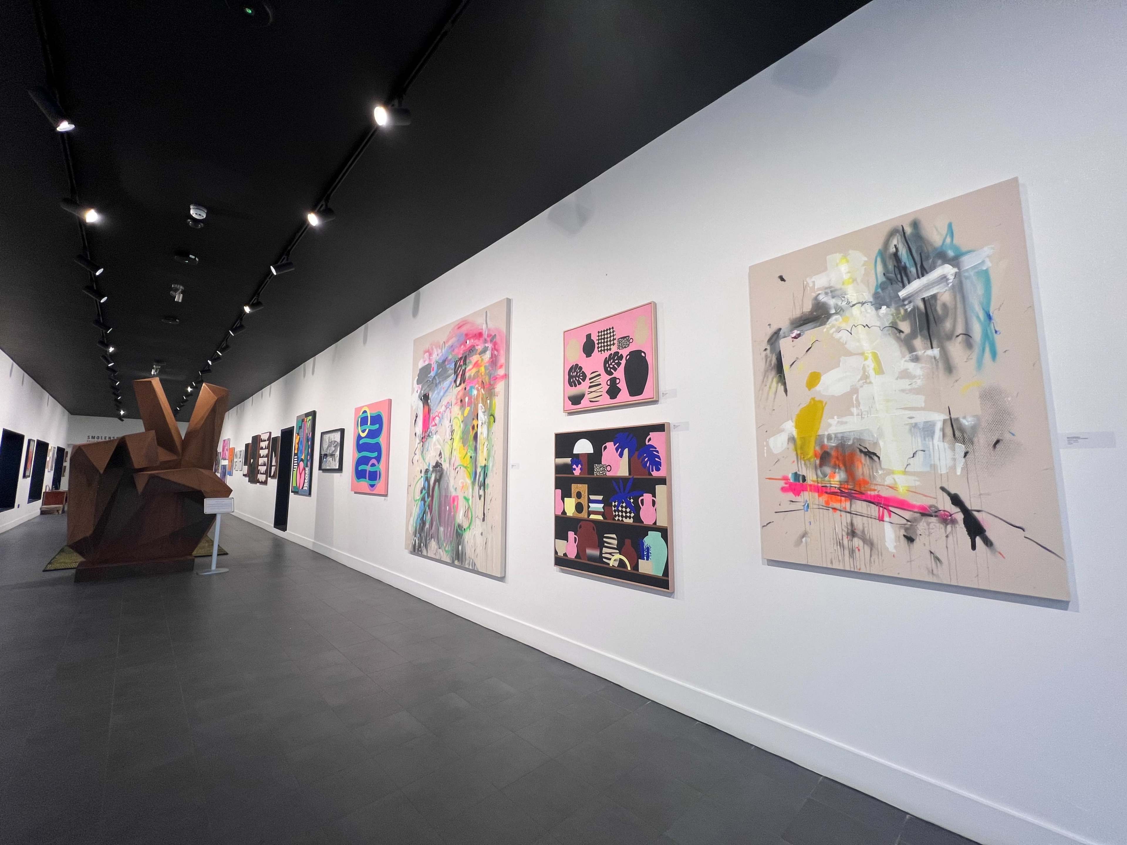 An art gallery interior with various colorful abstract paintings displayed on the white walls, alongside a large wooden sculpture.