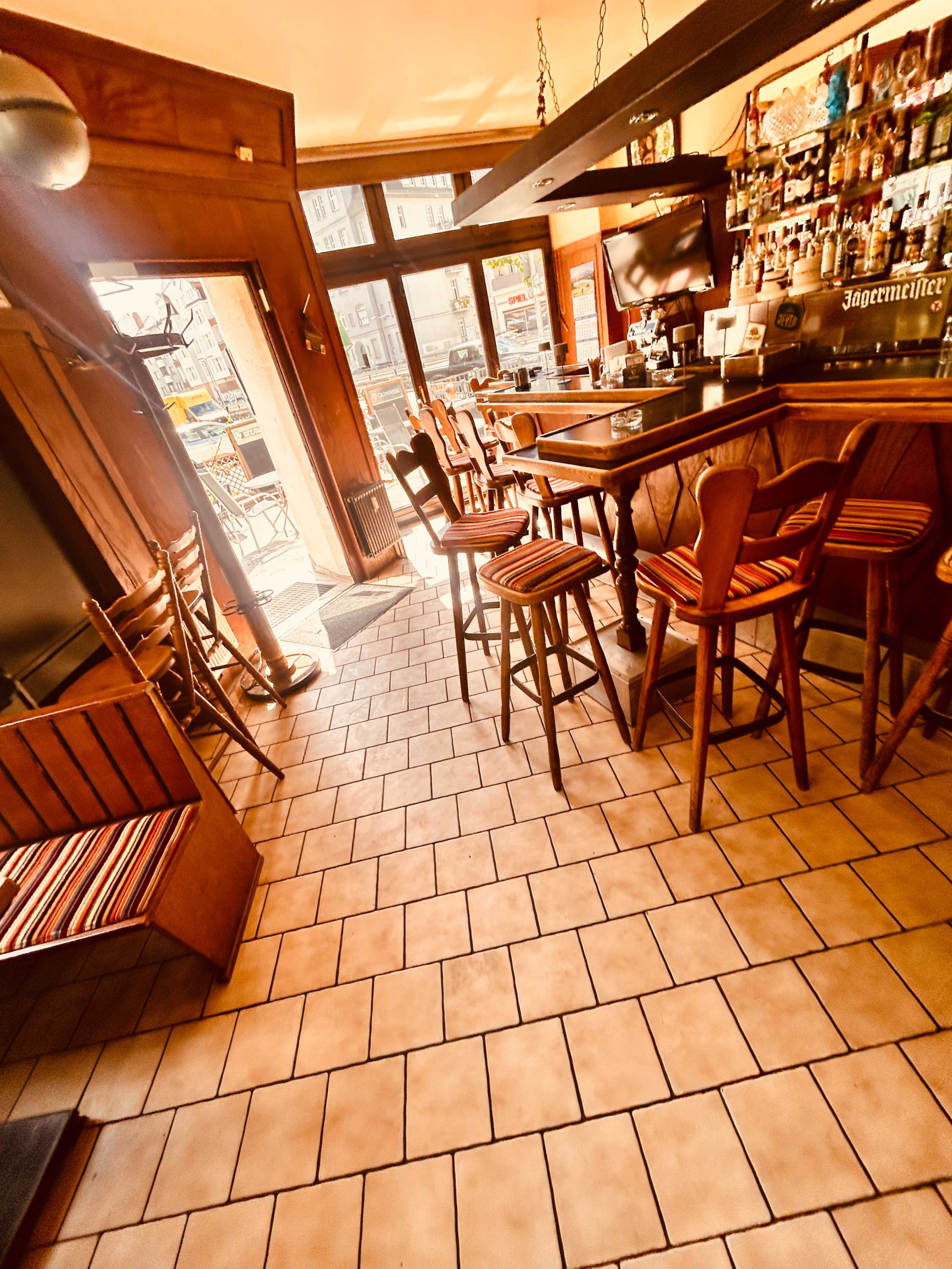 The image shows a bar area with wooden stools, tiled flooring, and a counter lined with various bottles behind it.