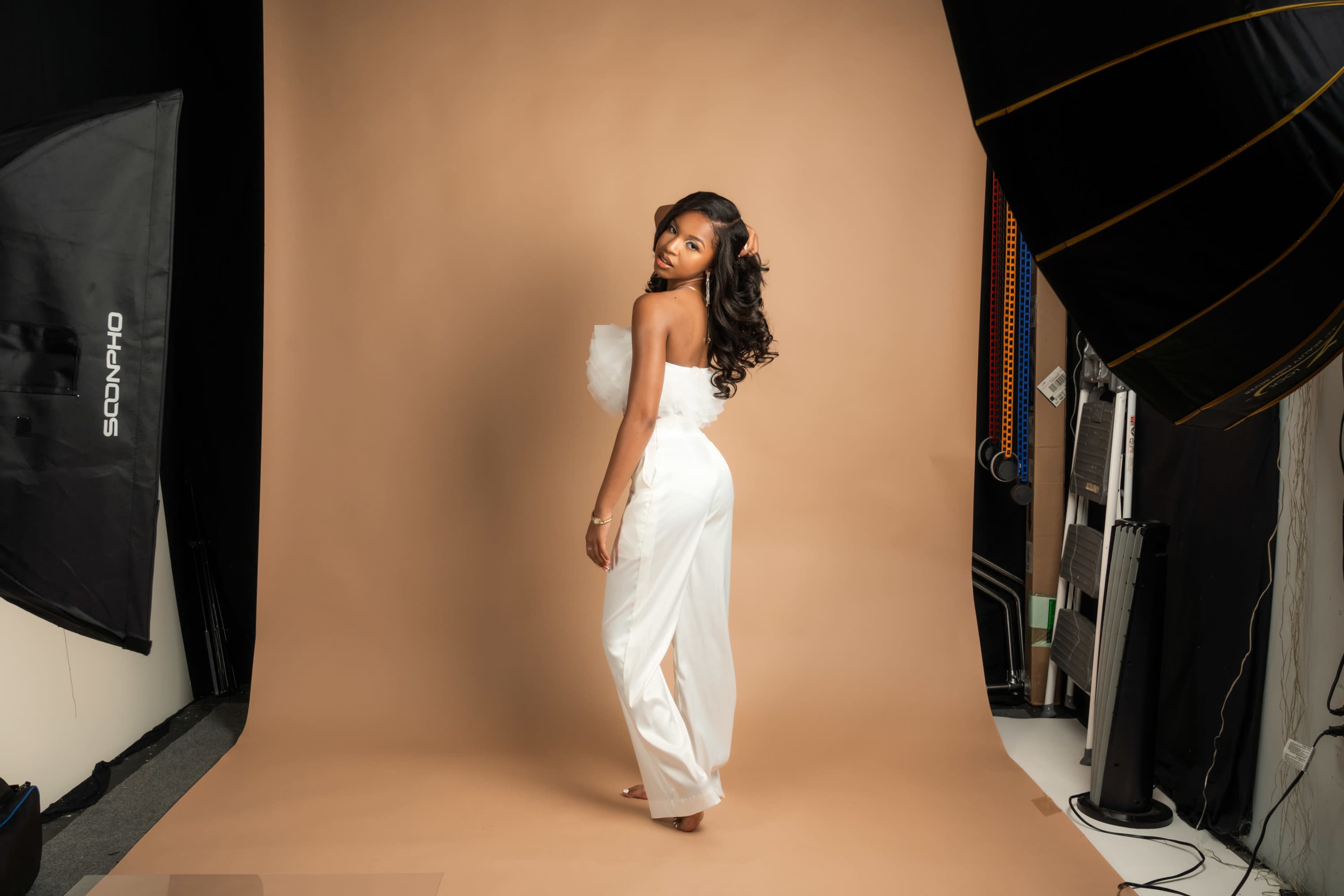 A woman in a white outfit stands against a light brown backdrop in a photography studio, surrounded by professional lighting equipment.