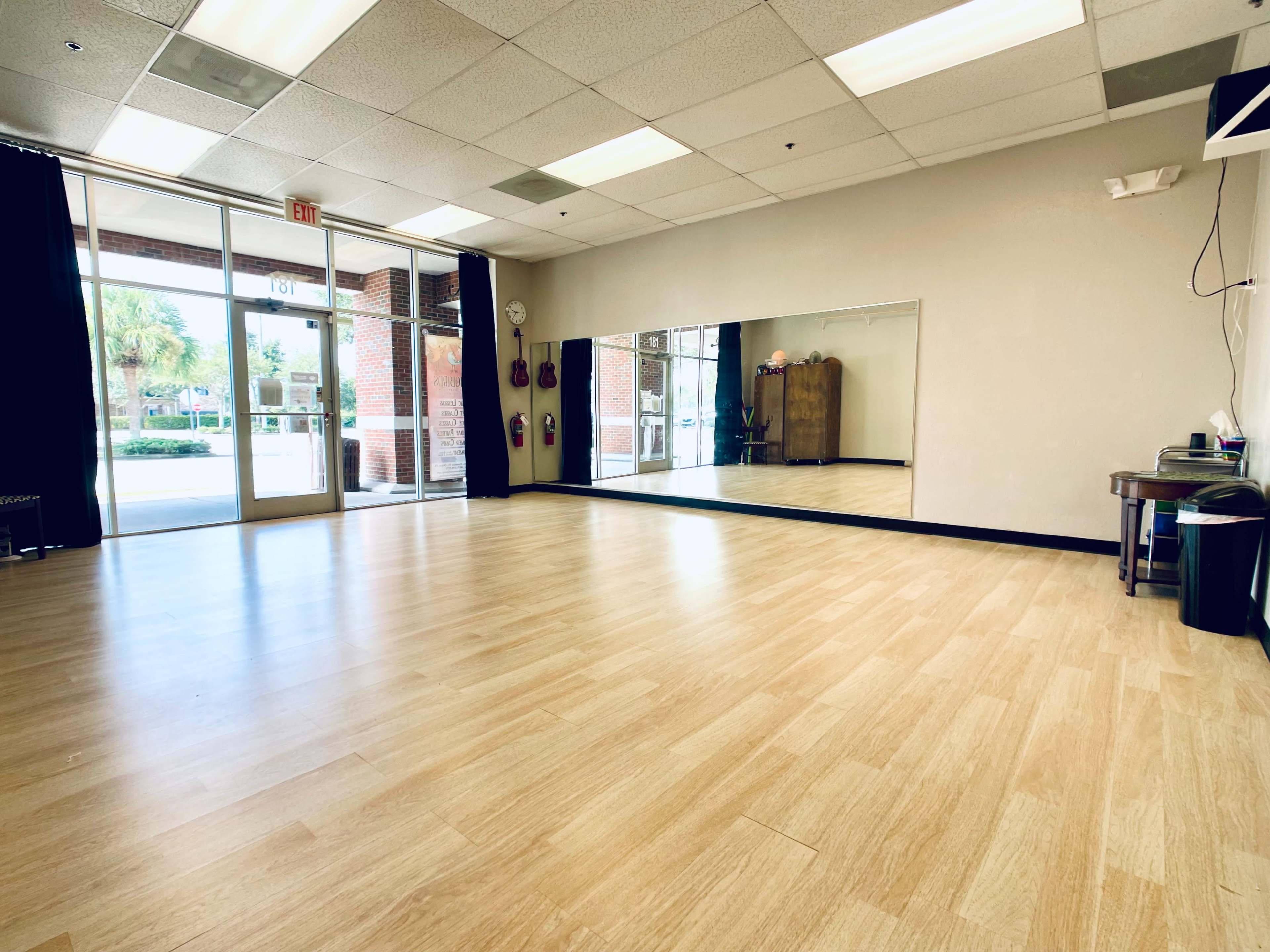 A spacious dance studio with wooden flooring, large mirrors on the wall, and natural light coming through the glass front doors.