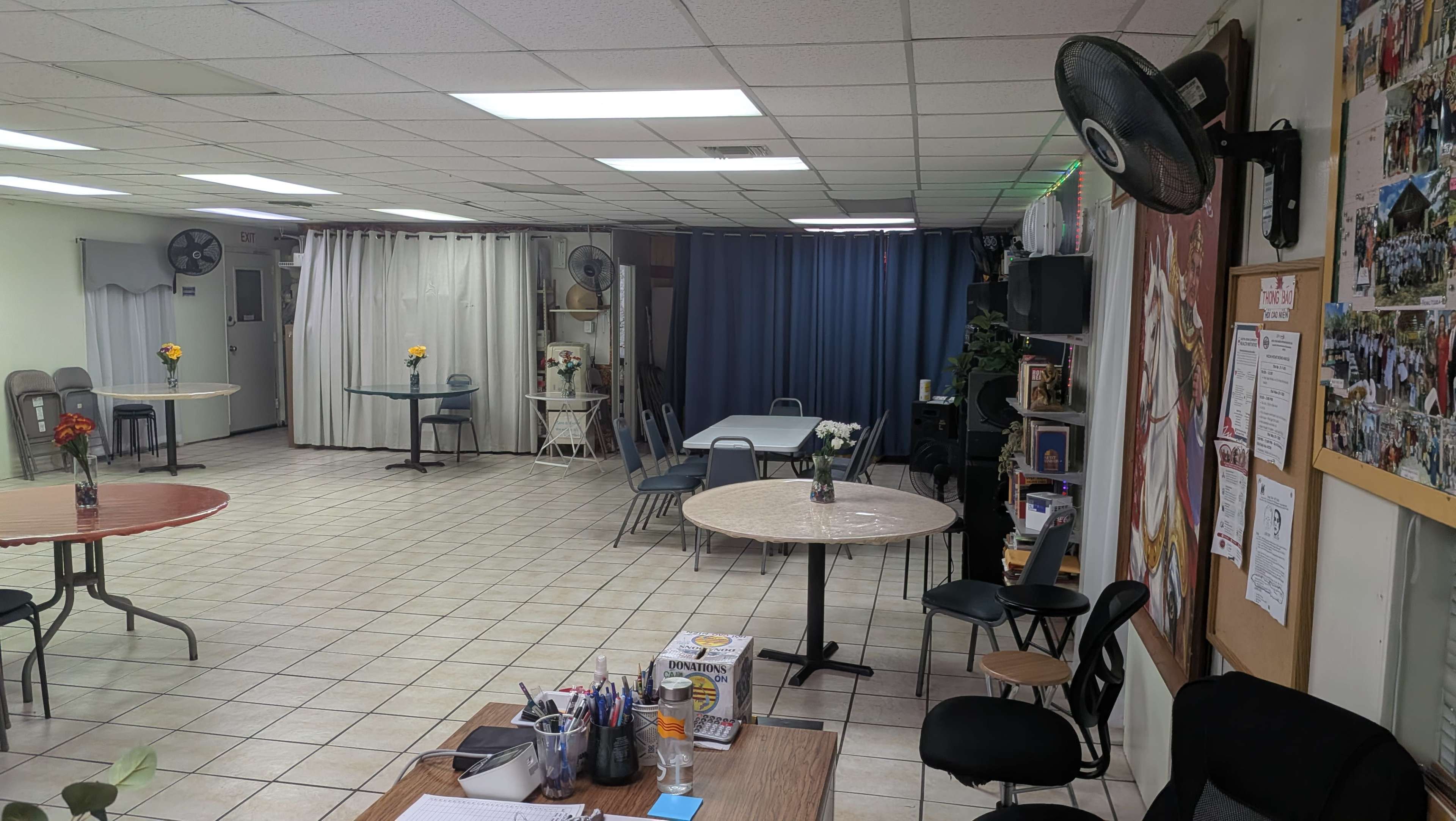 The image shows a spacious room with several tables and chairs arranged for use, featuring a small seating area, two fans, and decorative plants along the walls.