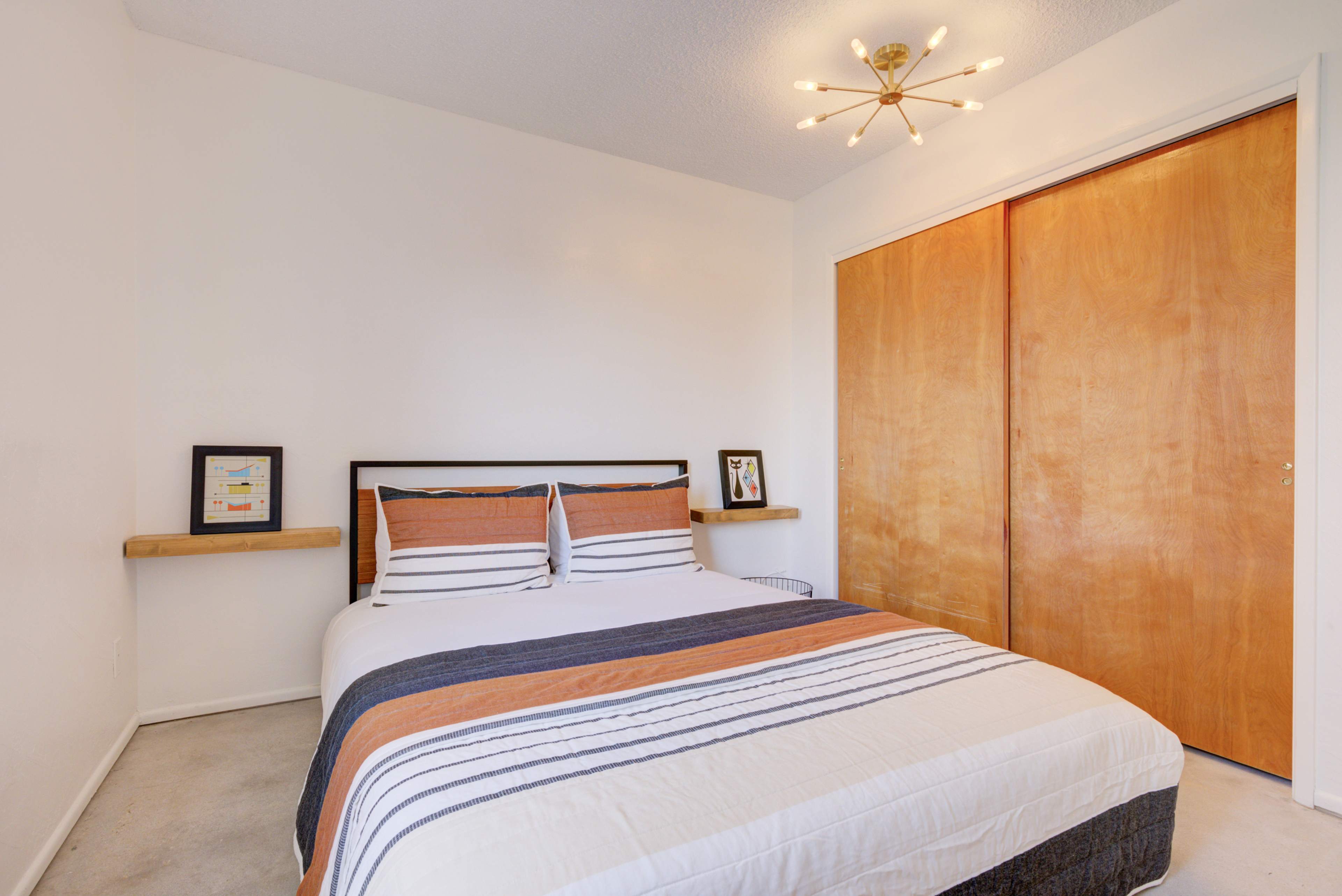 A minimalist bedroom with a double bed featuring striped bedding, two framed pictures on the wall, and a pair of wooden sliding closet doors.