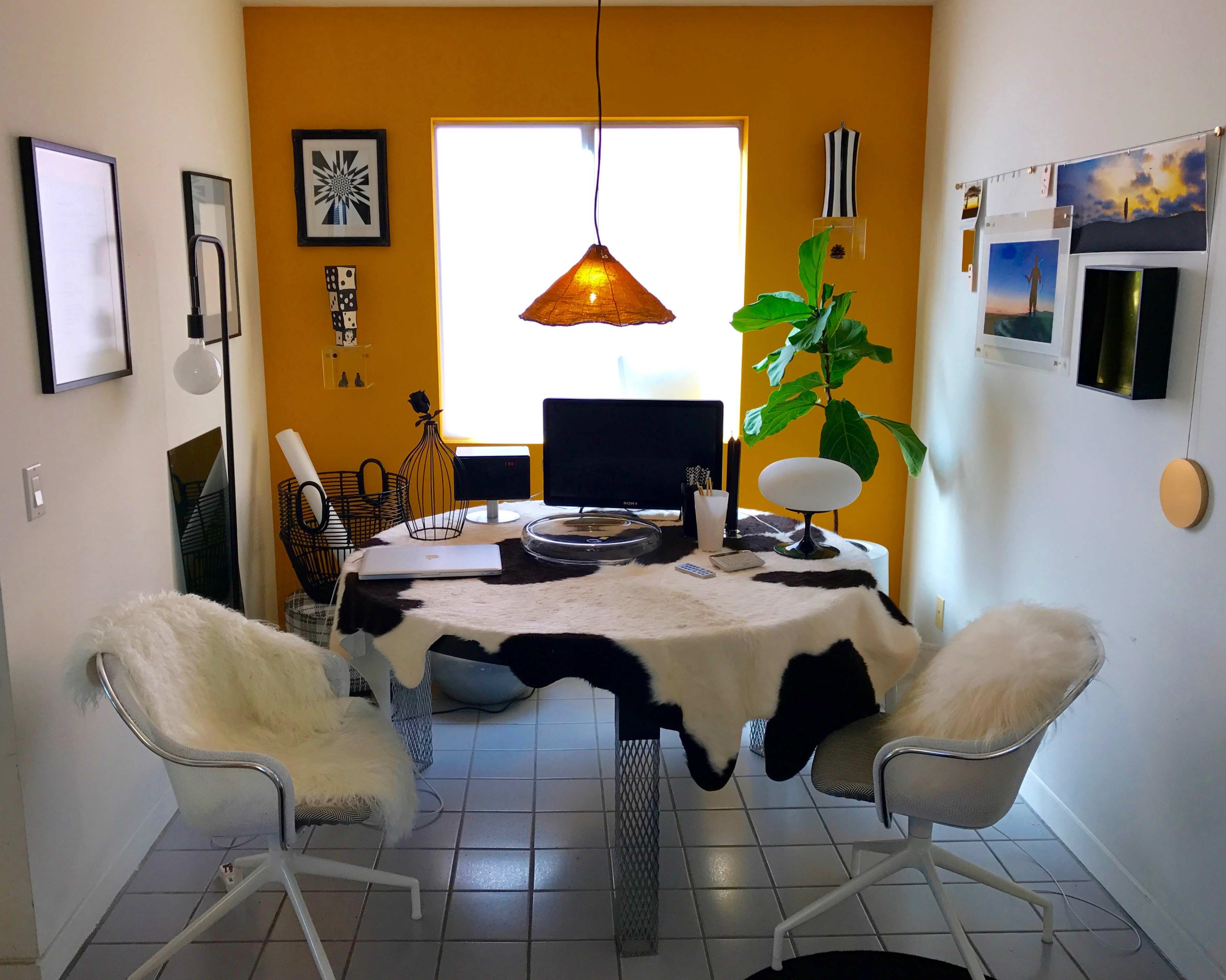 A minimalist home office features a round table covered with a cowhide rug, a computer setup, and a large window with yellow walls.