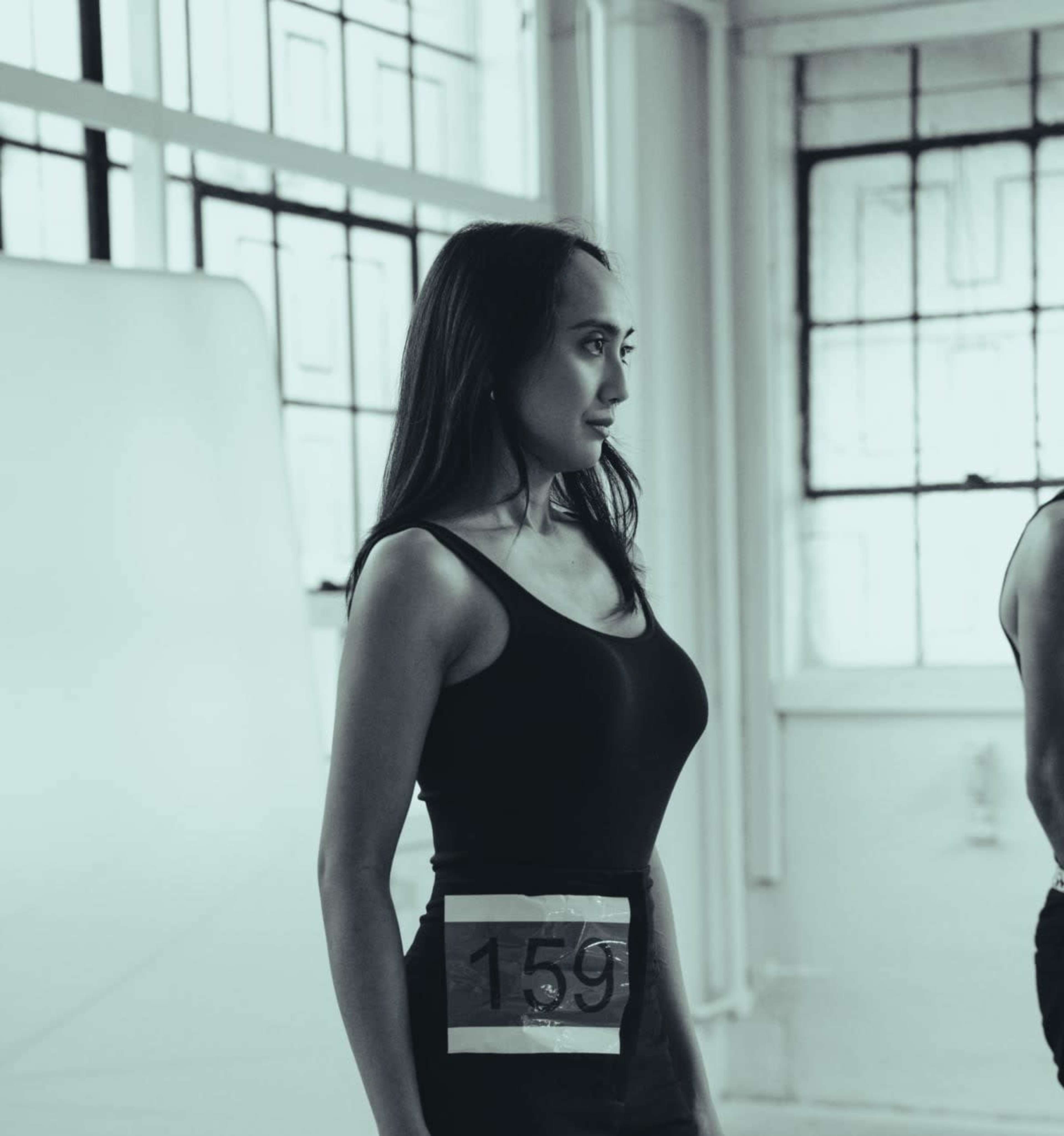 A woman stands in profile, wearing a black tank top and a number tag on her waist, during a casting or audition in a well-lit room with large windows.